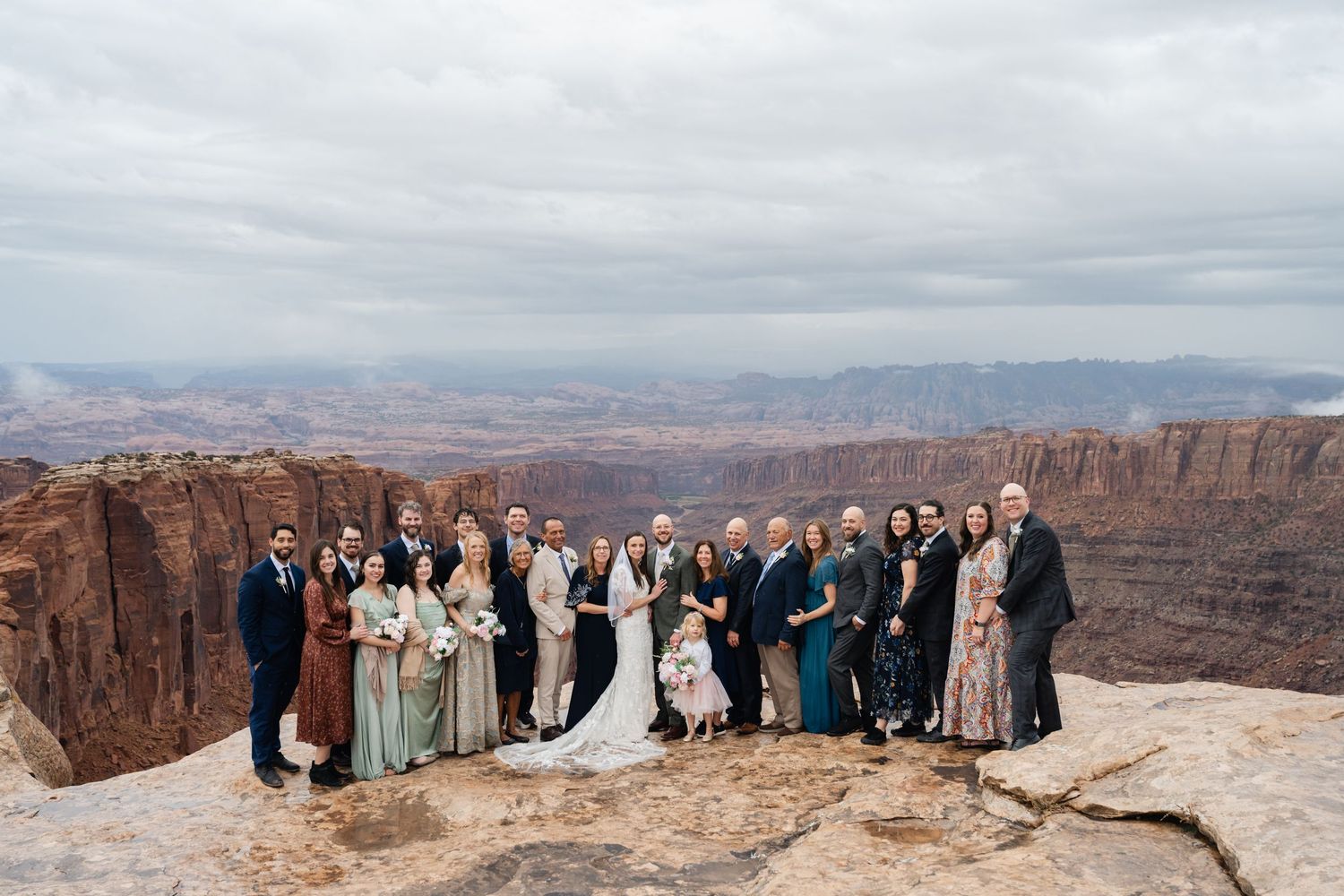 Panoramic shot of wedding celebration group spread across rocky outcrop with vast canyon views behind them.