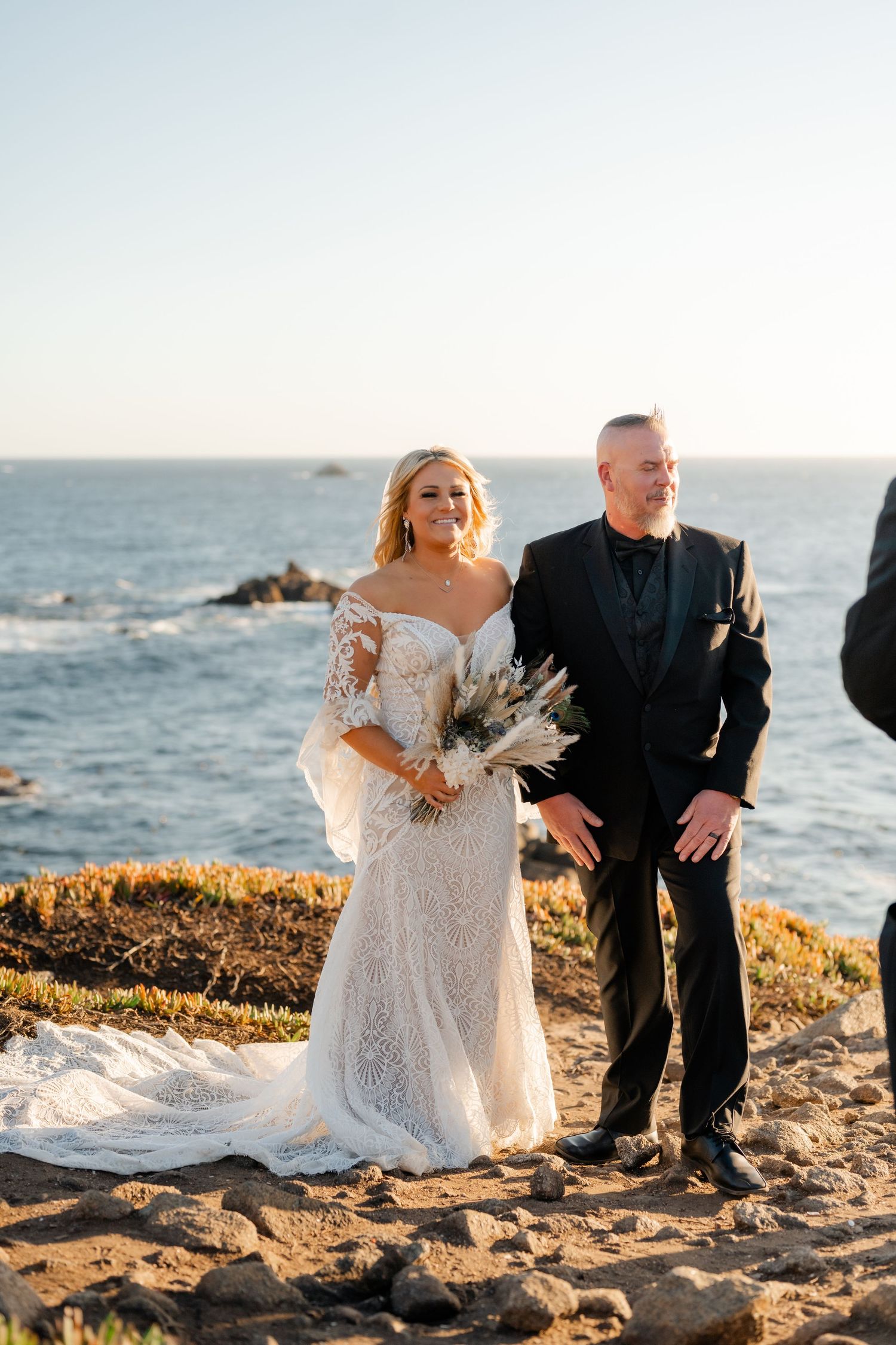 A romantic couple poses for wedding photos at sunset on a rocky coastal cliff overlooking the ocean.