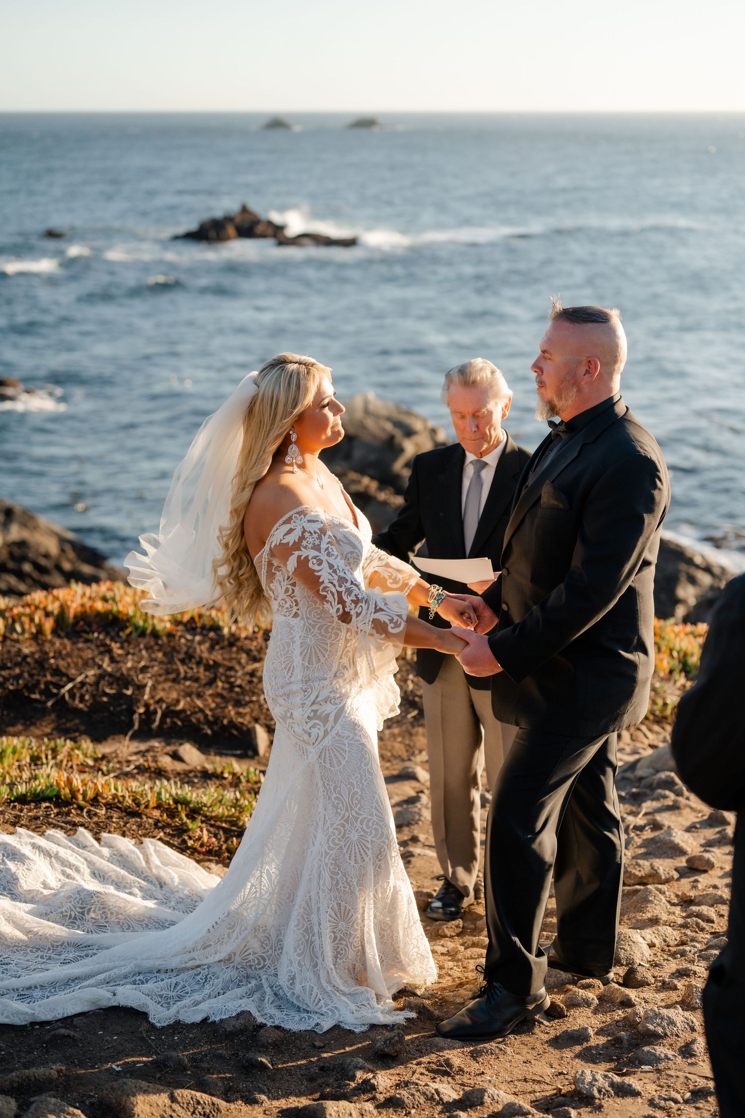 Wedding couple exchanges vows on scenic oceanfront cliff with officiant during golden hour.