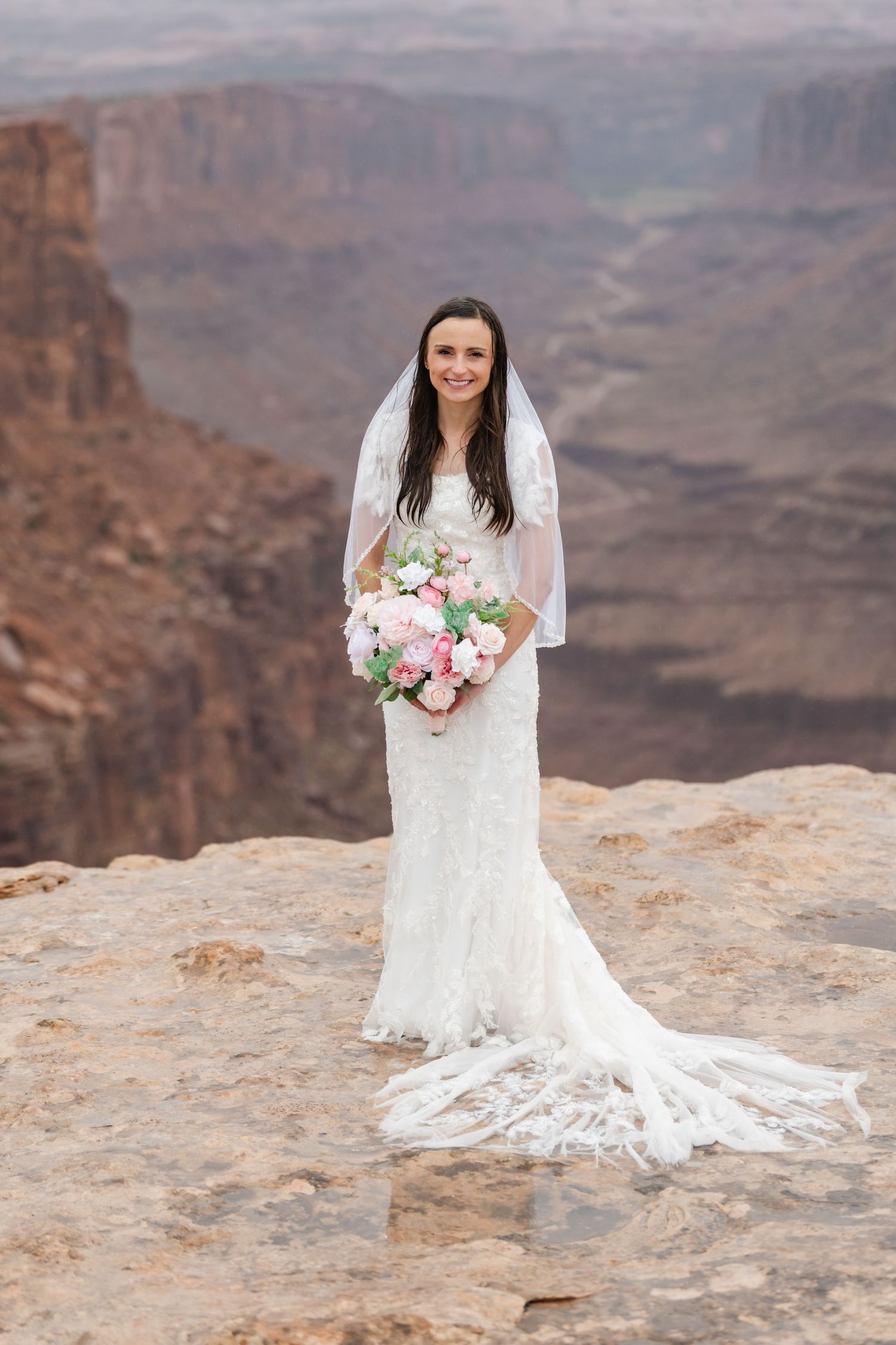 A bride in a lace wedding dress poses with a pink and white bouquet against the dramatic backdrop of the Grand Canyon.