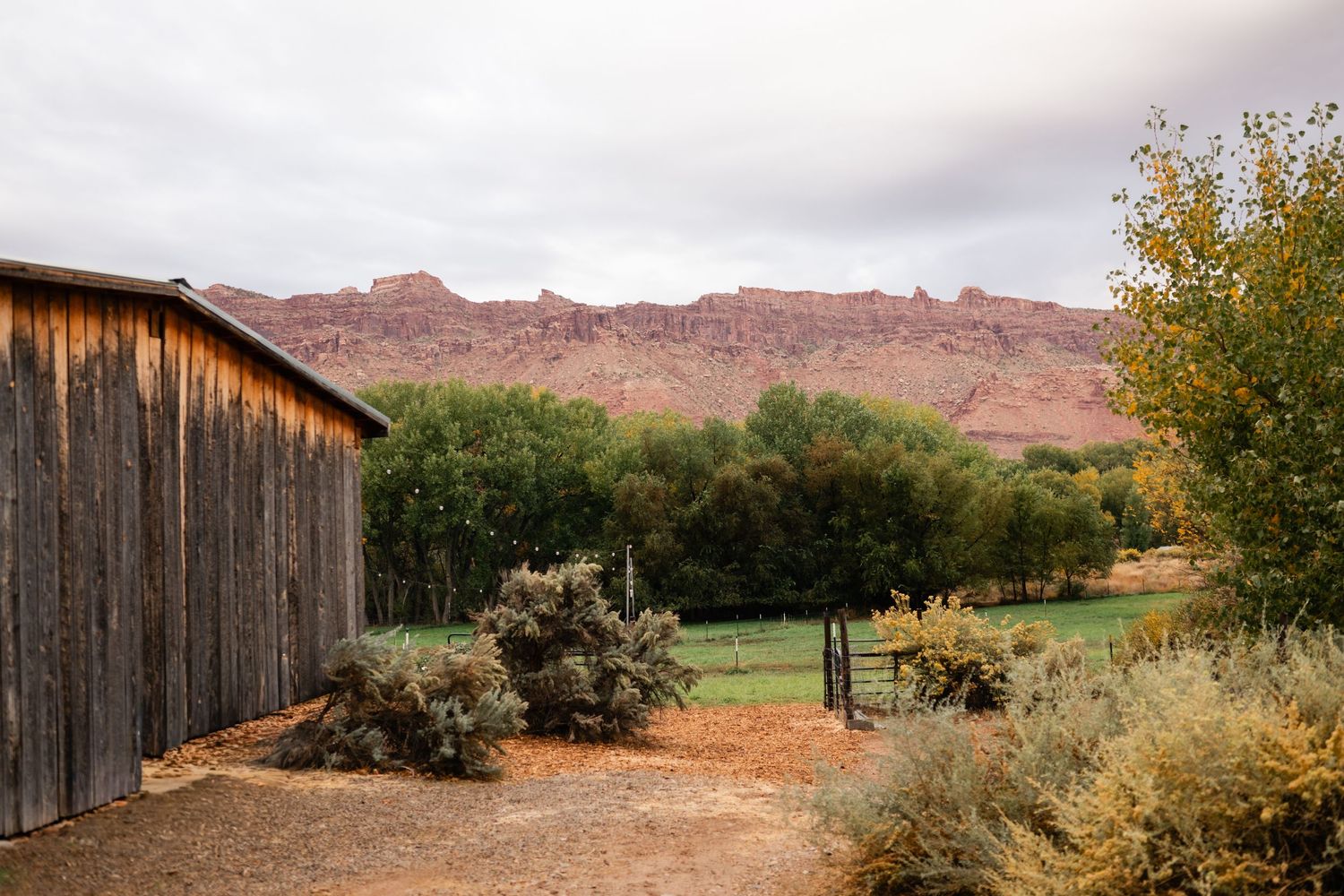 Rustic wooden barns and outbuildings stand against a backdrop of red rock cliffs in a desert landscape.