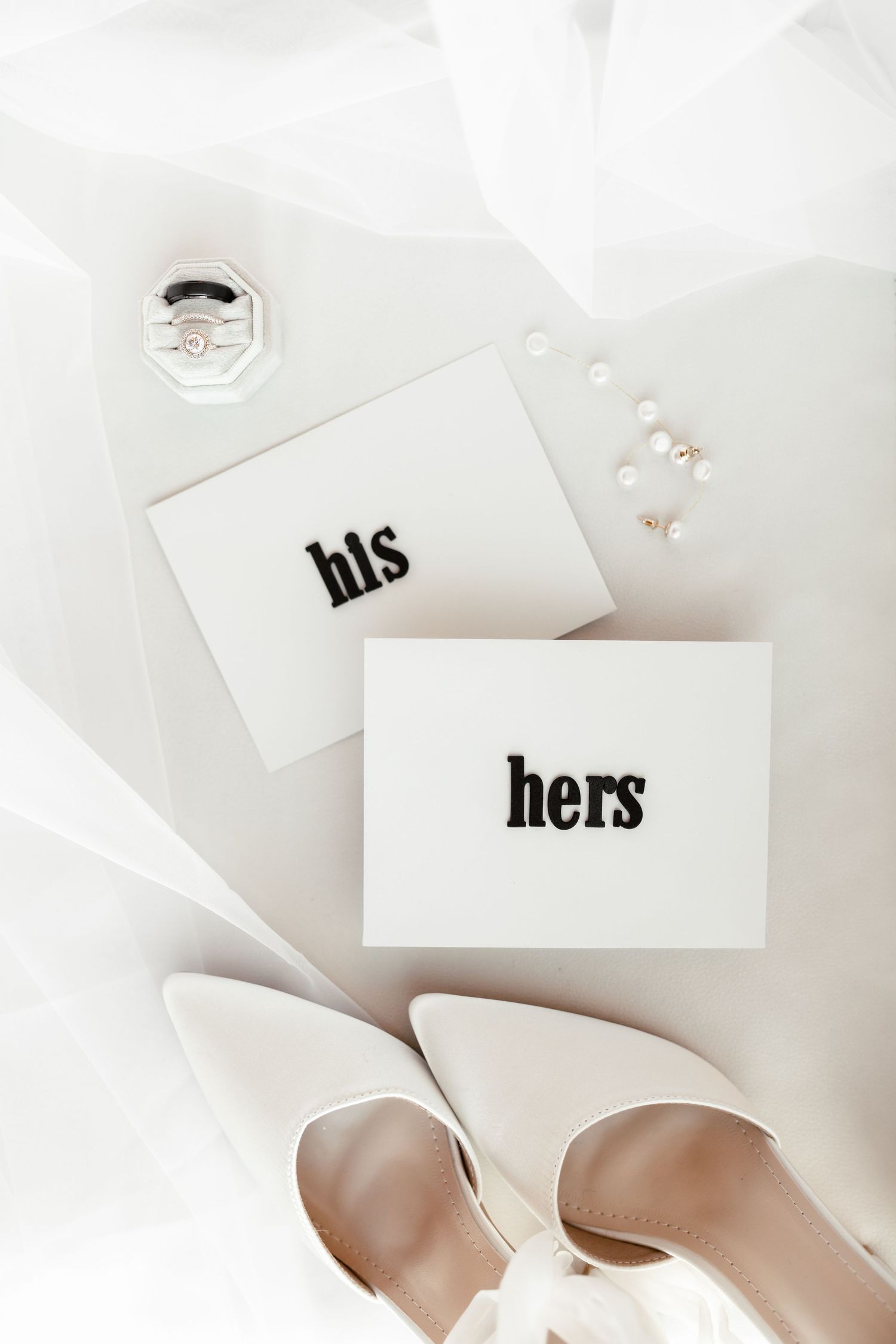 Wedding stationery with 'his' and 'hers' cards and delicate jewelry on white background.