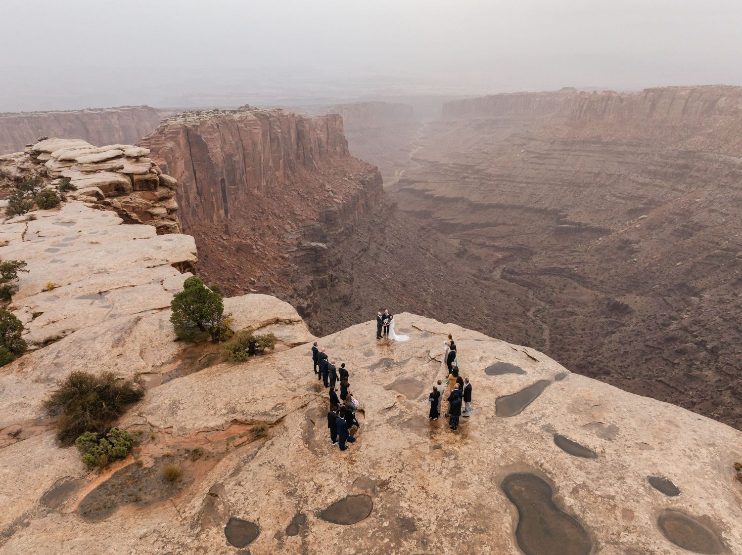 Visitors stand at the edge of a vast desert canyon overlook with red rock formations stretching to the horizon. Moab Wedding Photographer.