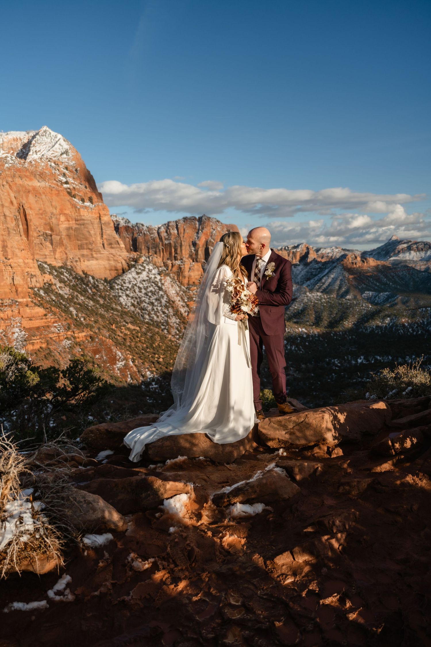 Wedding portrait on red rock overlook with snow-capped canyon peaks in background.