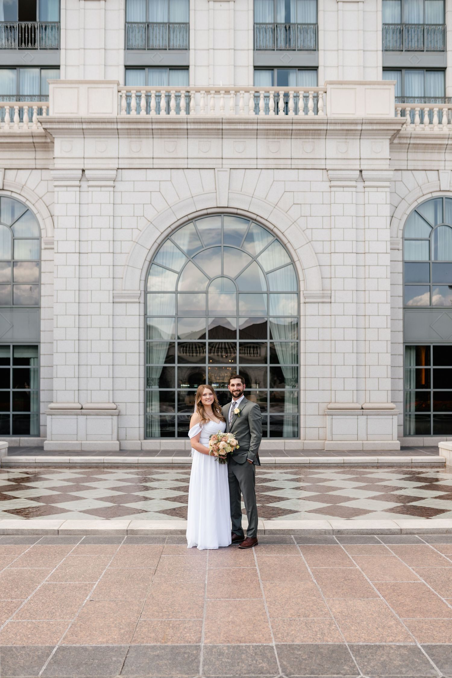 Elegant wedding couple poses against white stone building facade with arched windows and balustrade details.