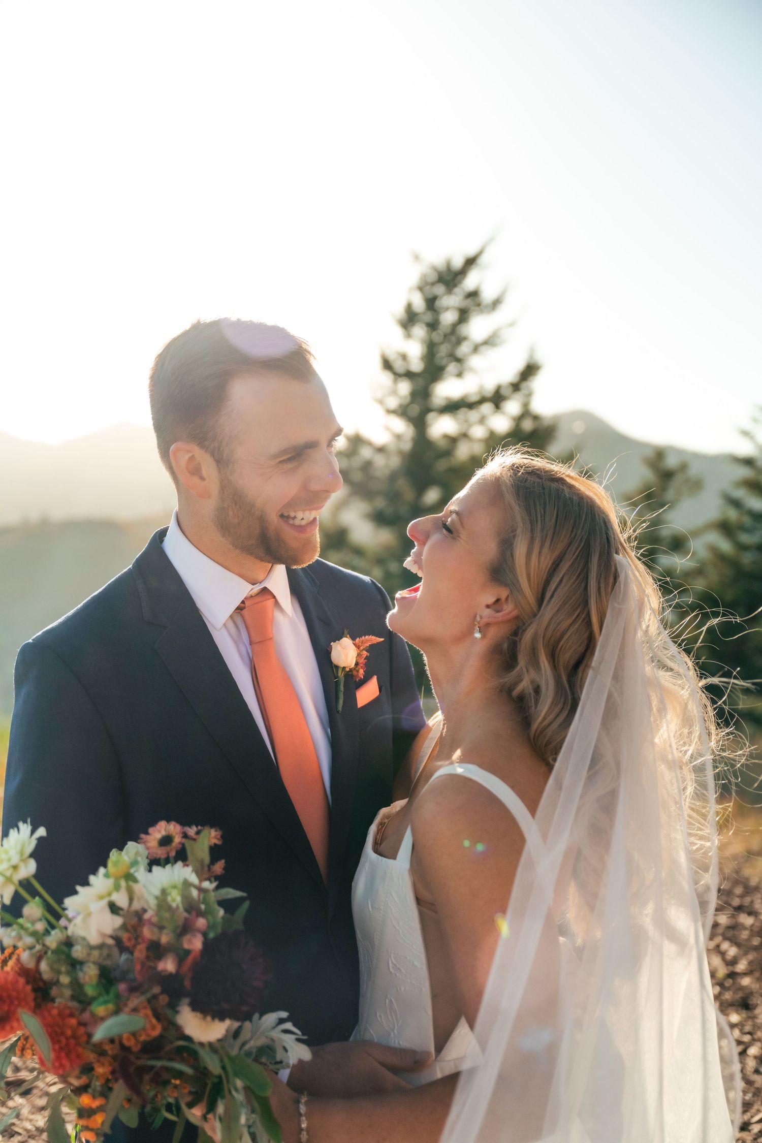 A joyful wedding couple shares a candid moment during golden hour with coral and white floral bouquet.