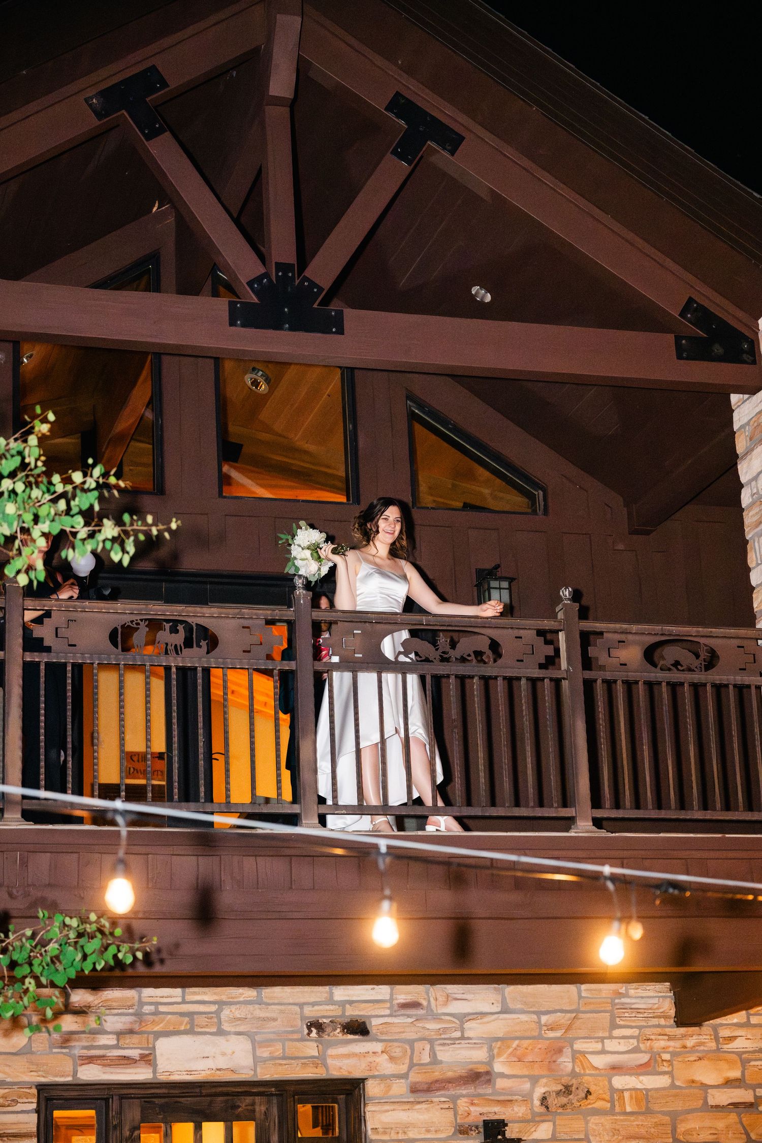 Bride in white dress poses on rustic balcony with wooden beams and stone walls in warm lighting.