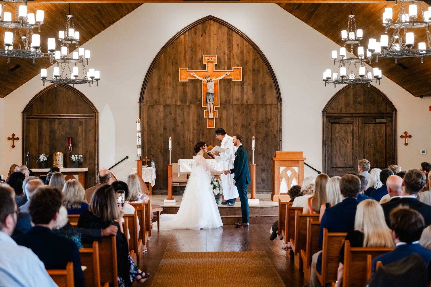 Wedding ceremony in progress inside a chapel with wooden altar and chandeliers.