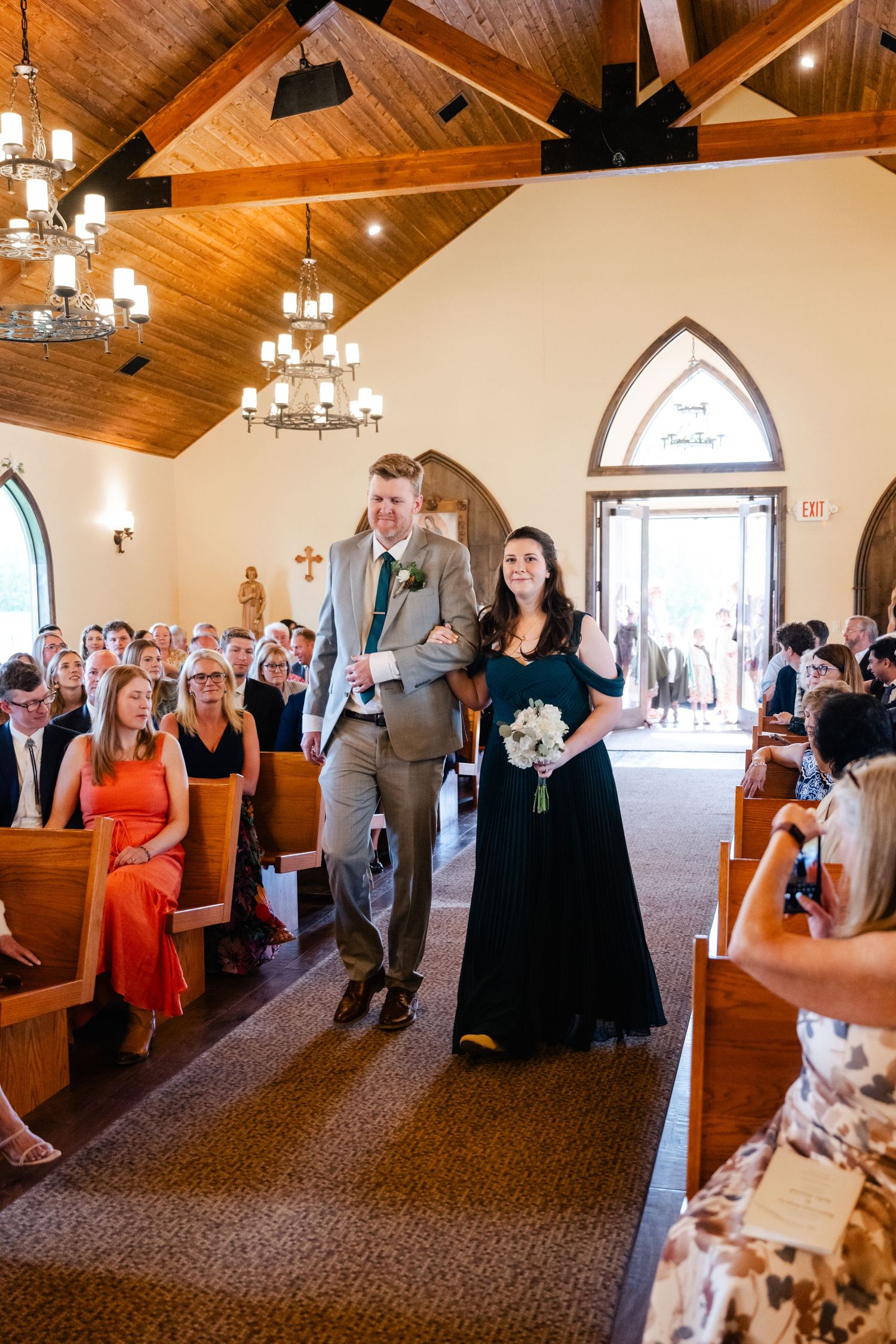 Wedding guests stand in a church sanctuary with wooden beams and arched windows during a ceremony.