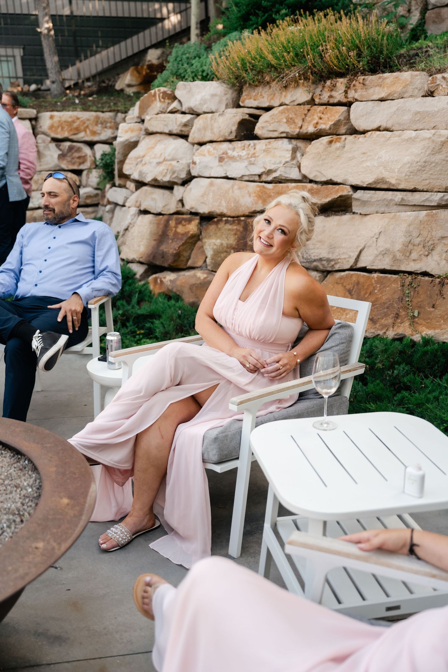 A person in a pink dress relaxes in white Adirondack chairs against a stone wall backdrop.