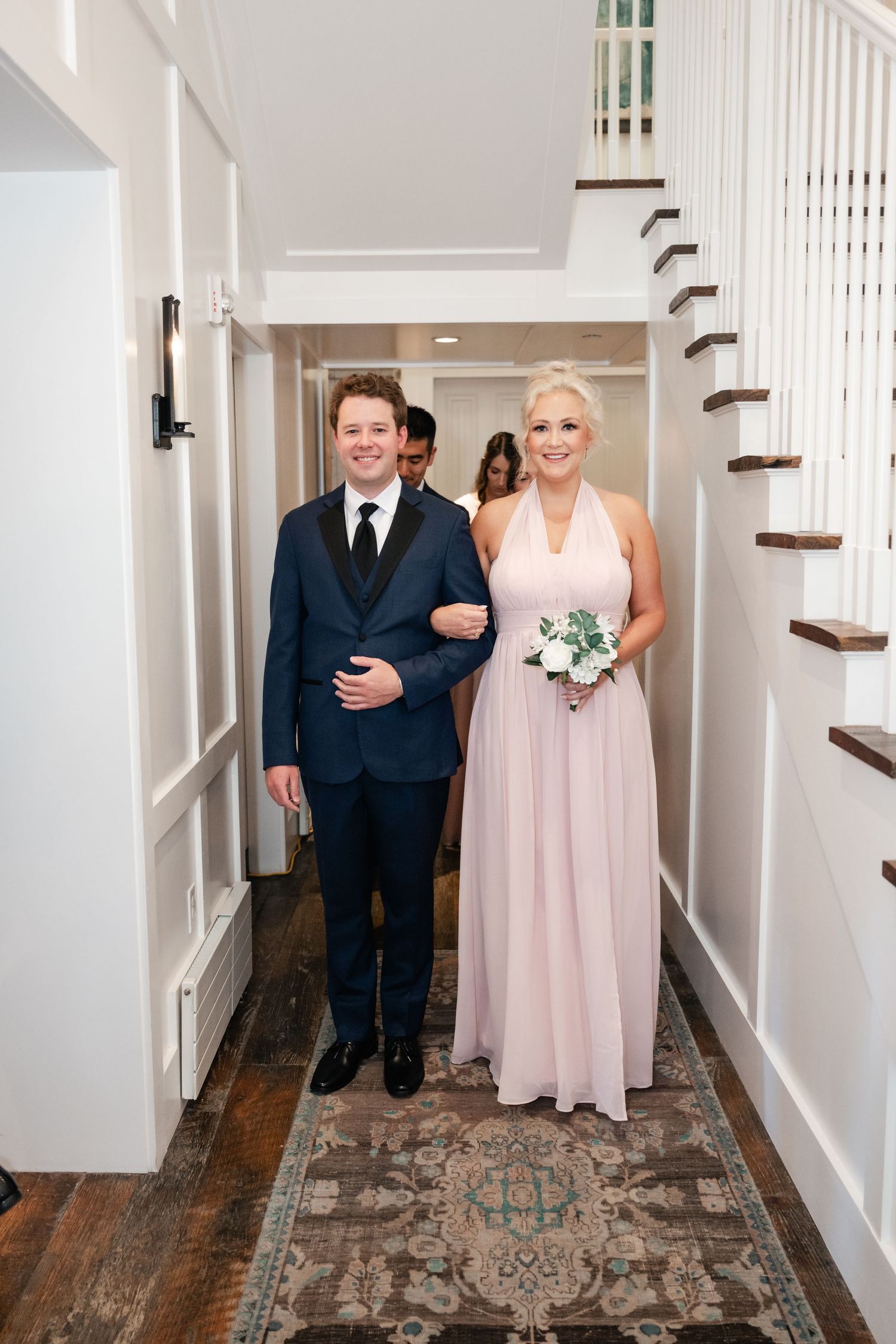 Wedding party members walking down a hallway with white wainscoting and hardwood floors.