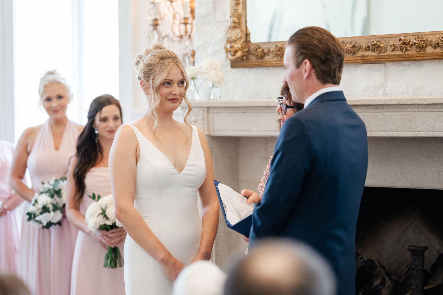 A couple exchanges vows during an indoor wedding ceremony with bridesmaids in pink dresses standing nearby.