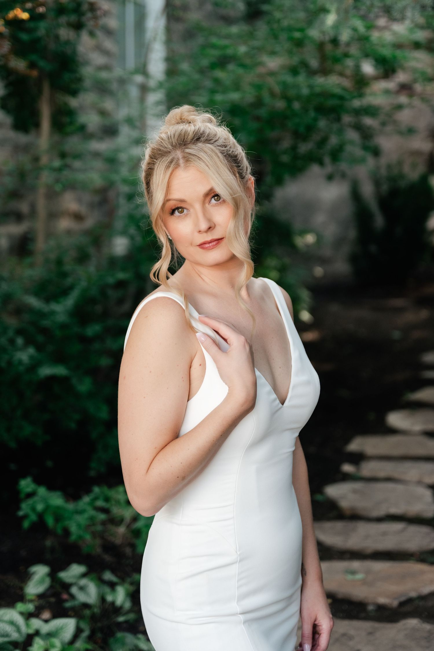 Elegant bride in a white wedding gown poses in a garden setting with stone walls.