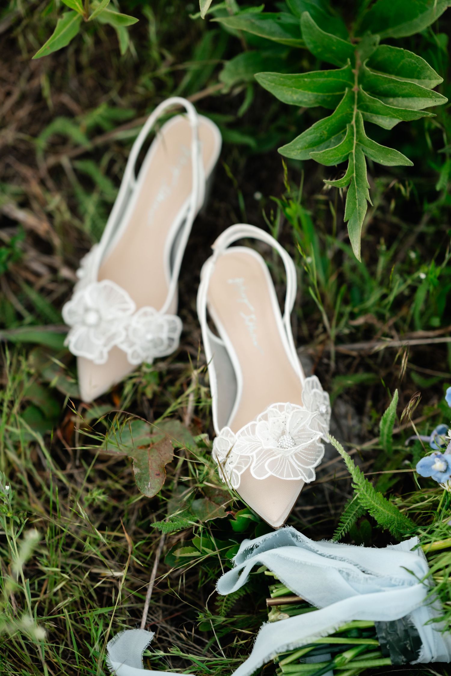 Elegant white bridal shoes with lace details placed among green foliage.
