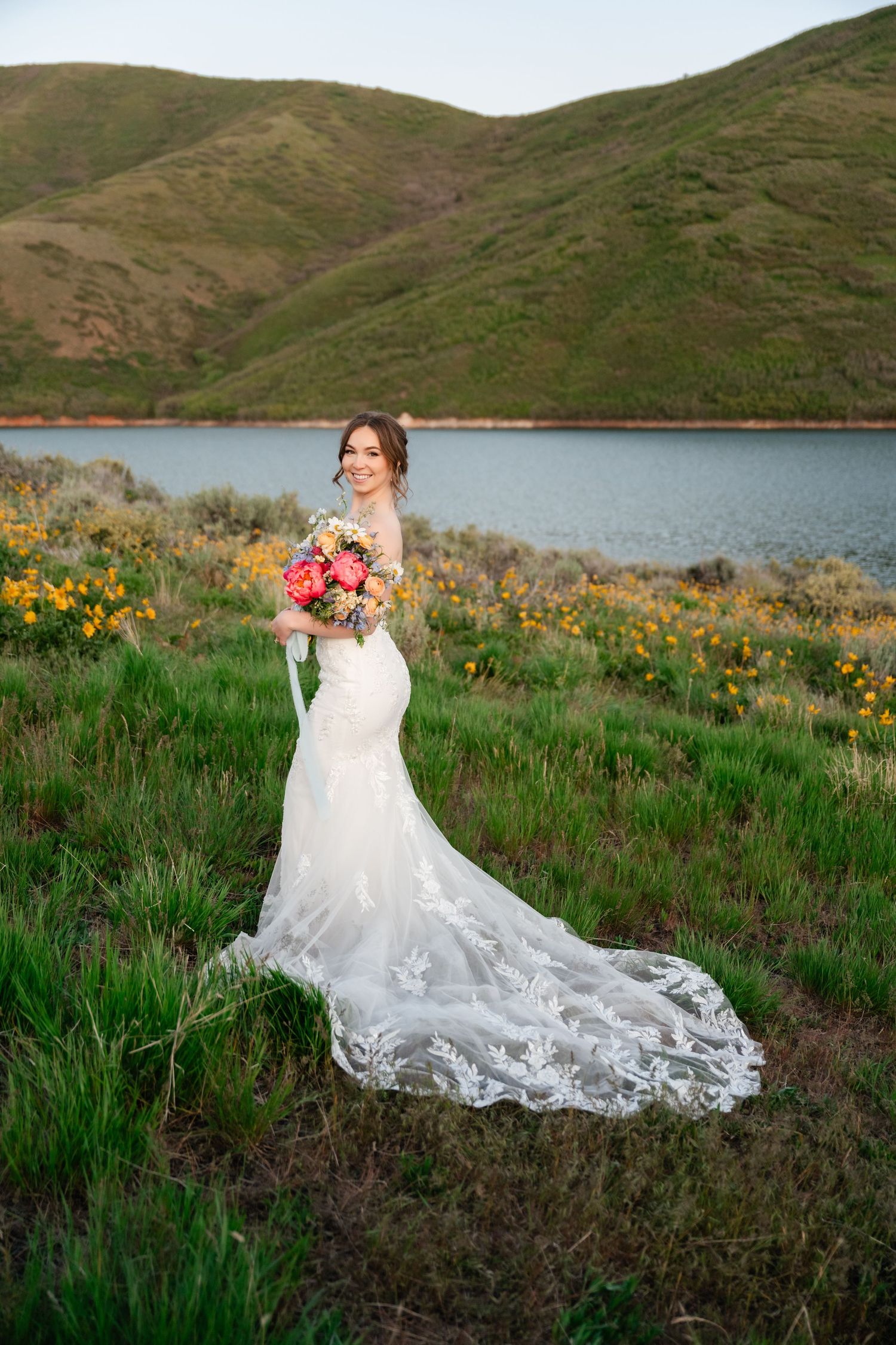 Bride in lace wedding gown with long train poses against green hills and mountain lake backdrop.