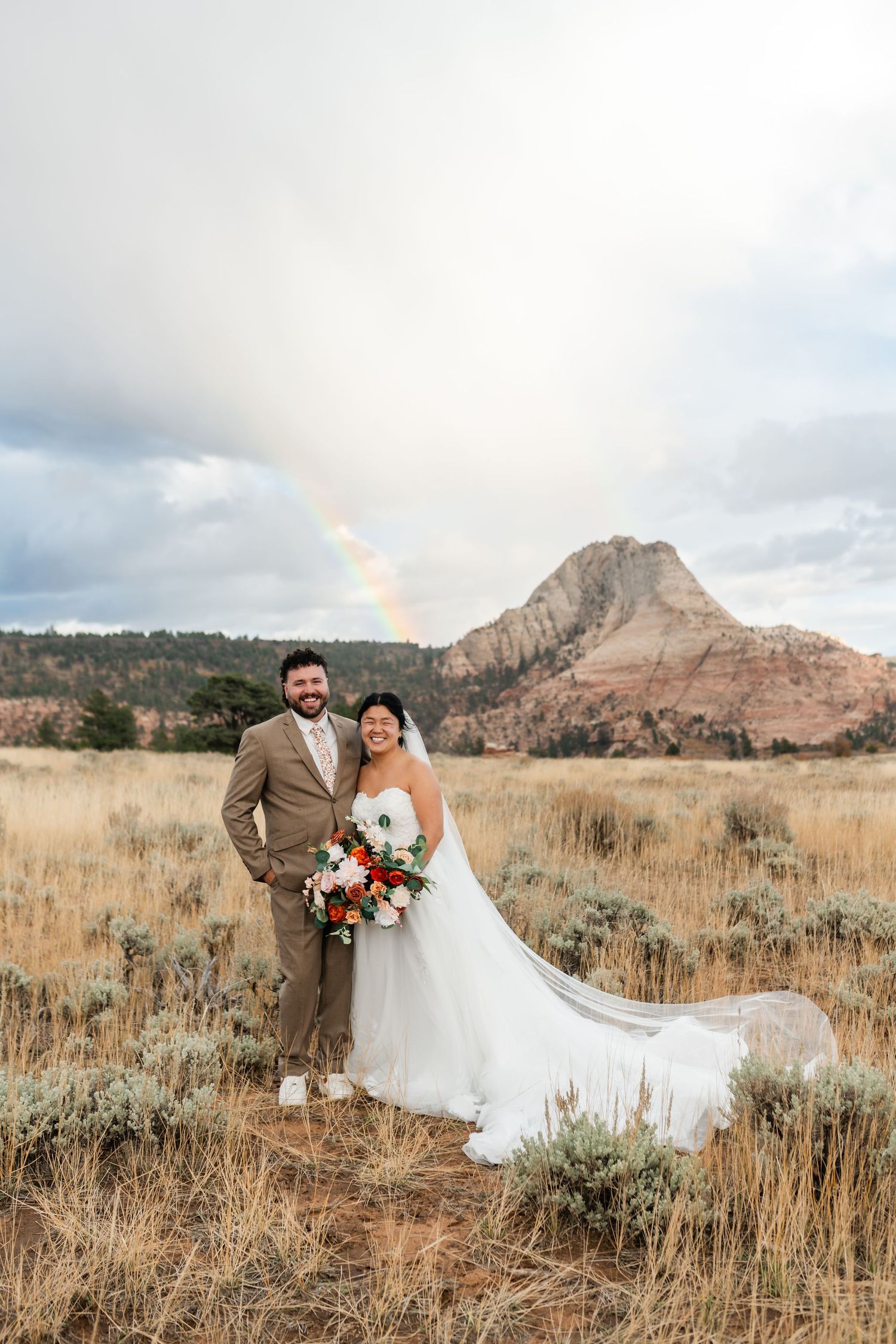 A wedding couple poses in a desert field with mountain peaks and cloudy skies.