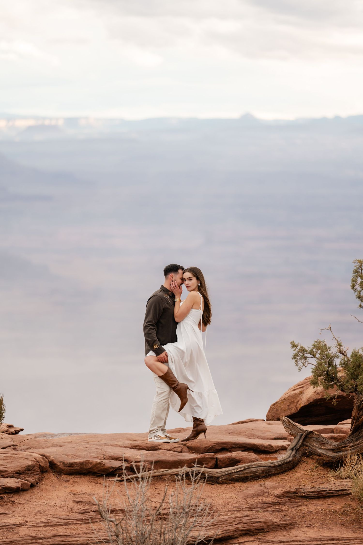 A couple embraces on red rocks overlooking a misty desert canyon landscape at sunset.