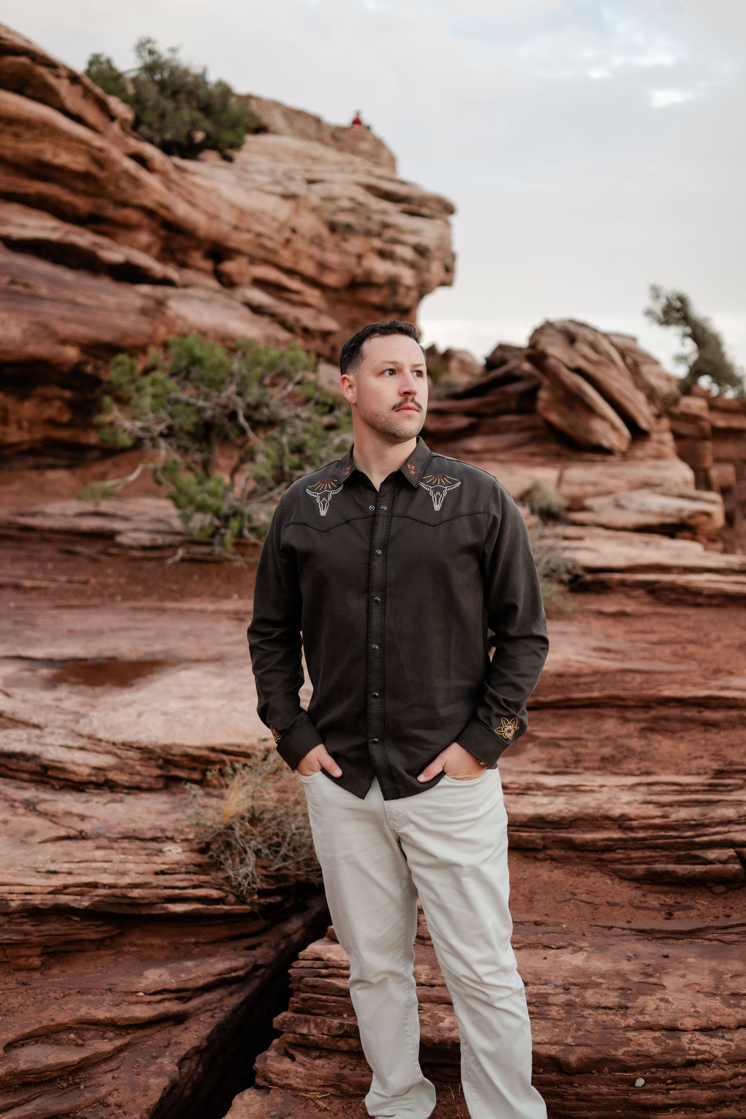 Person in dark button-down shirt and light pants standing against dramatic red rock formations.