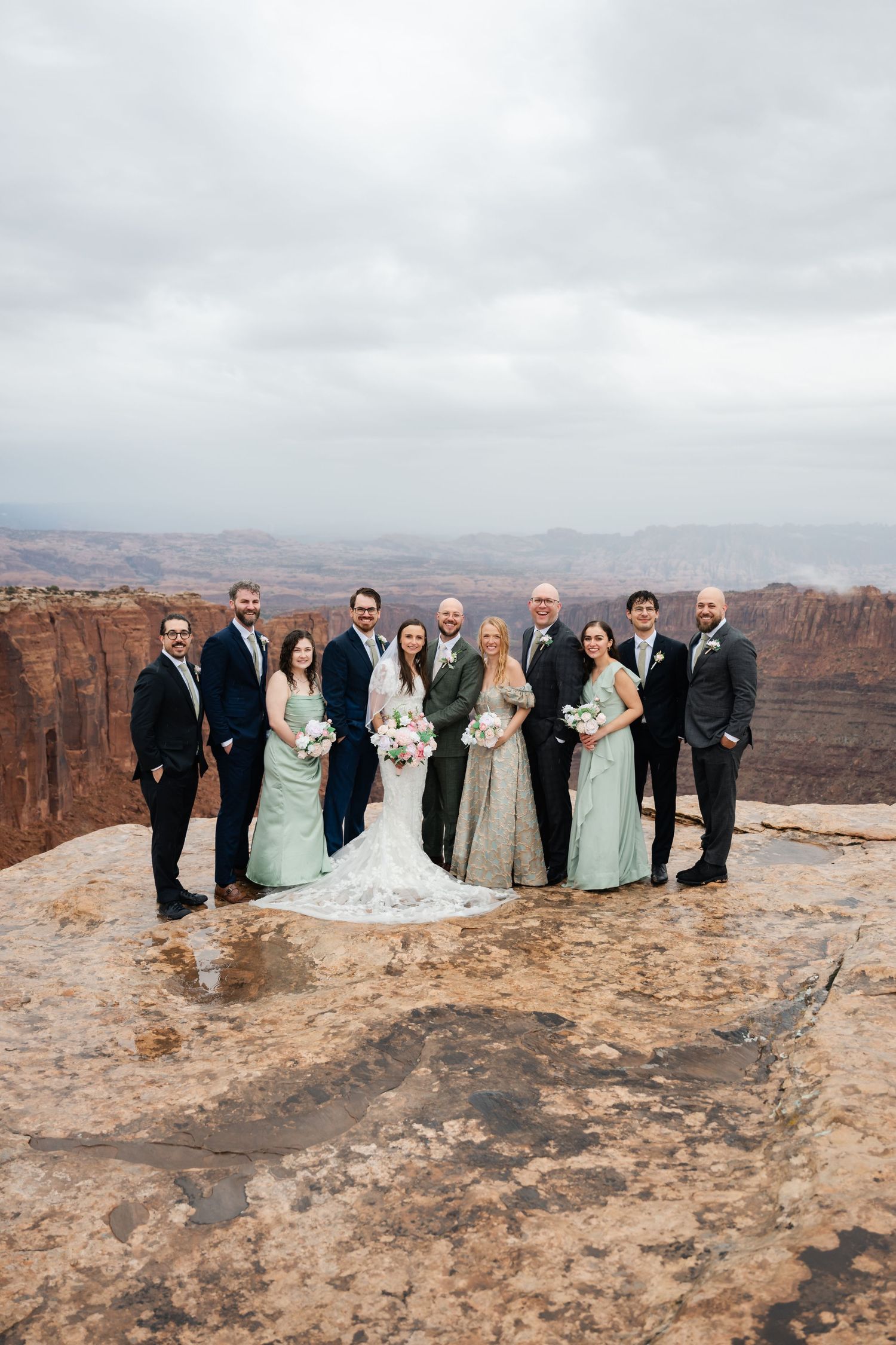 Wedding party stands together on a dramatic cliff edge overlooking the vast canyons of Moab, Utah. Moab Wedding Photographer.