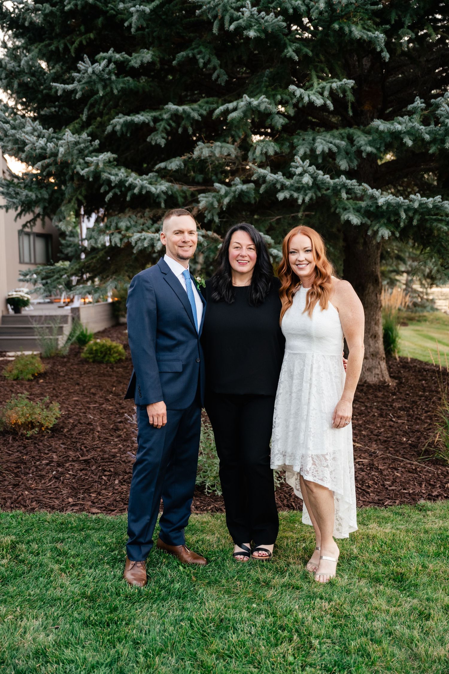 Group photo of wedding party members standing together on grass near evergreen trees.