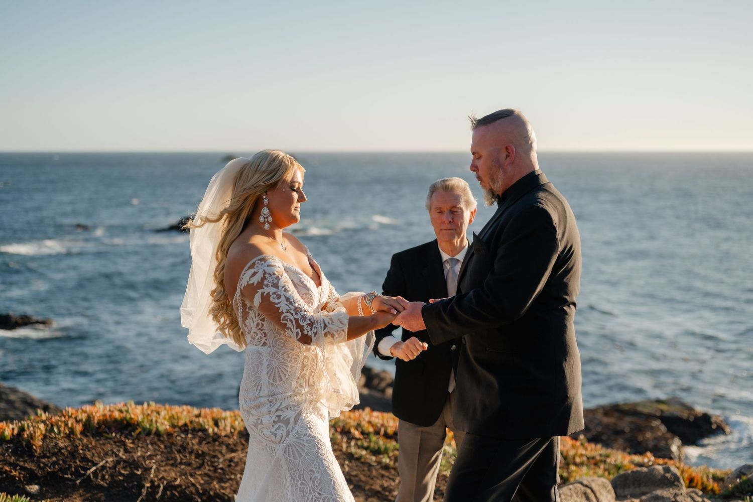 An intimate seaside wedding ceremony unfolds against a backdrop of blue ocean waters and rocky coastline.