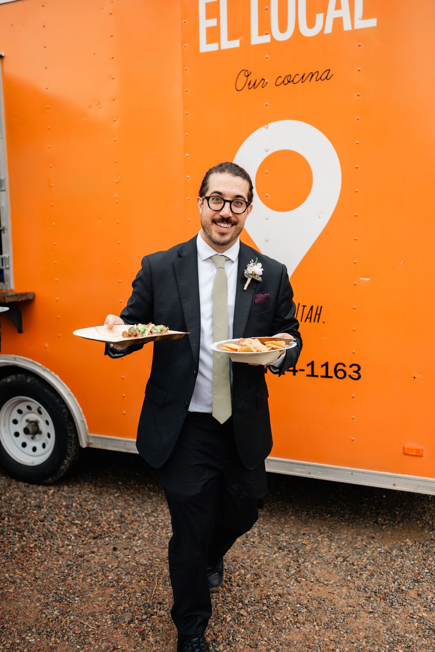 Server in formal attire carrying plates of food in front of an orange food truck labeled 'El Local'.