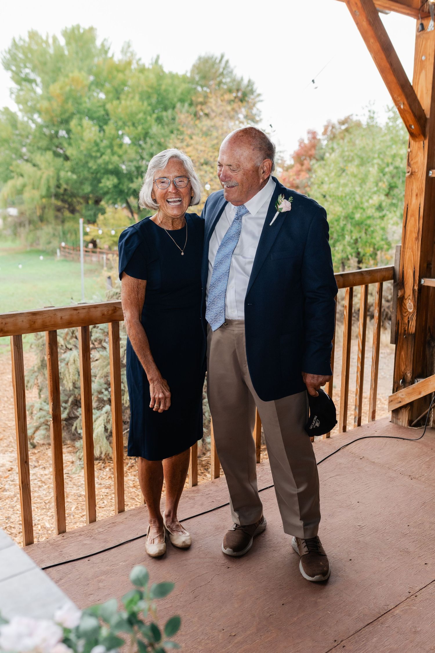 A couple in formal attire standing together on a wooden deck with autumn trees in the background.