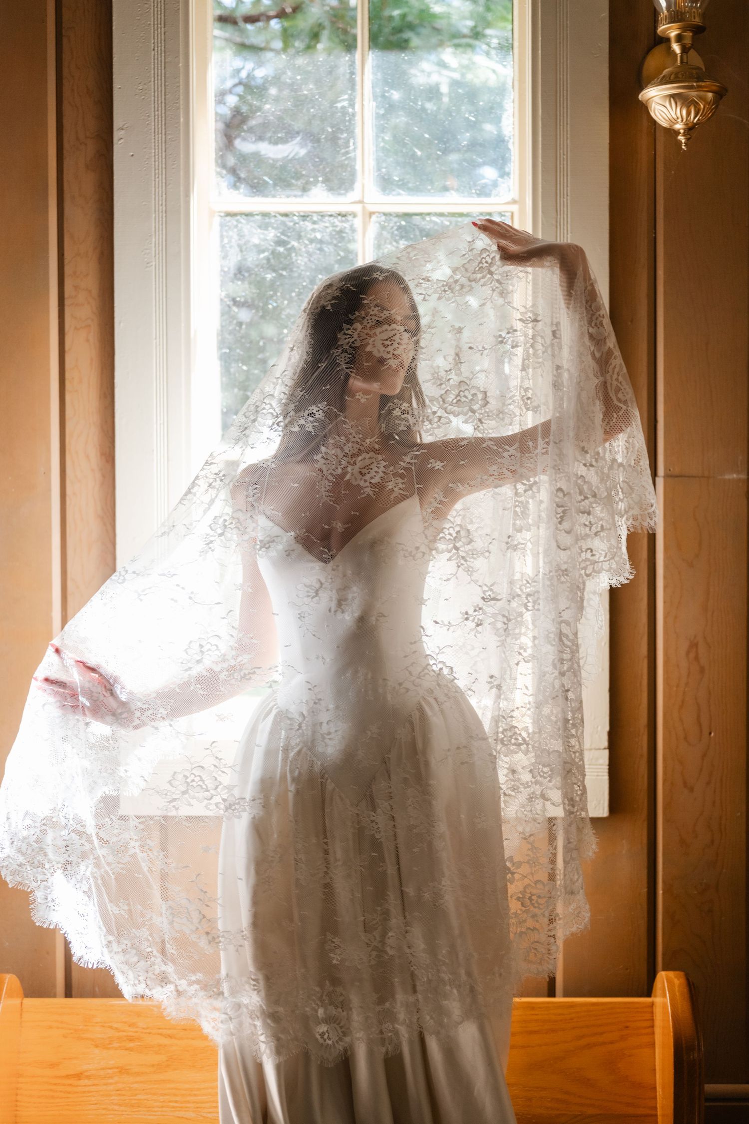 Bride in lace wedding dress and veil poses gracefully by a sunlit window in a warm wooden interior.