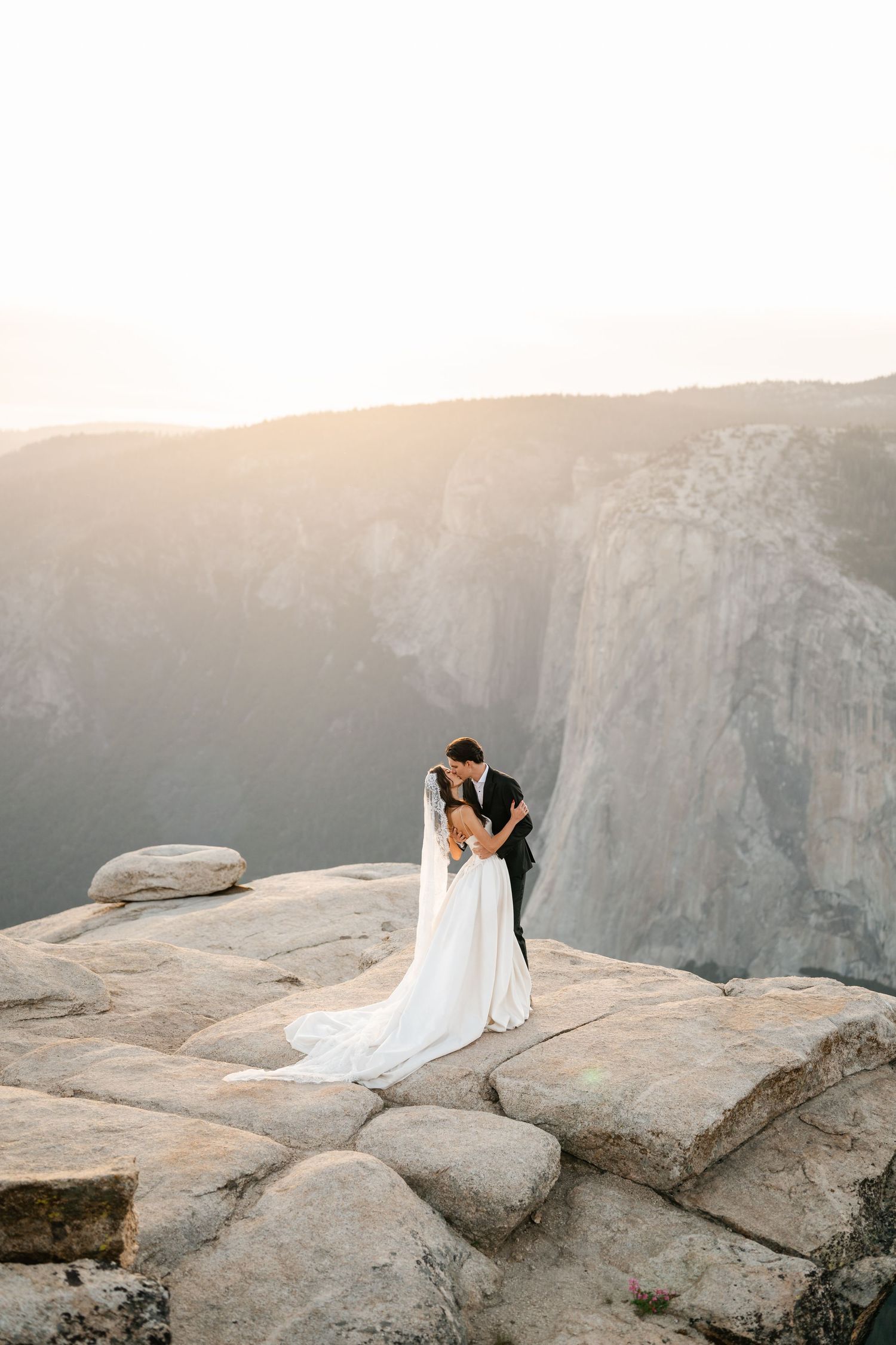 Bride and groom share romantic embrace on rocky mountain overlook at sunset with valley views behind.