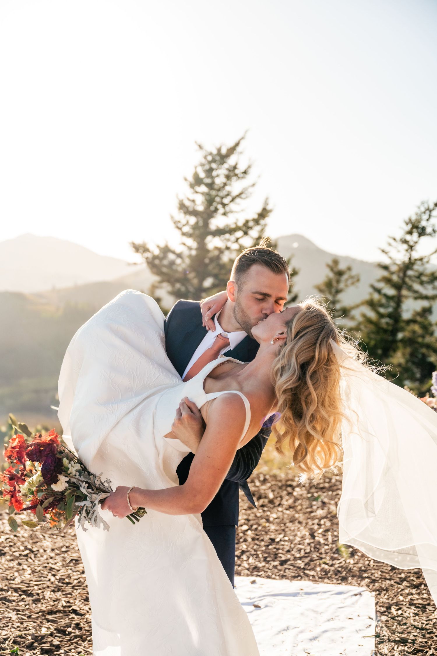 Romantic wedding couple embraces in a sunset mountain setting with evergreen trees and a flowing bridal veil. Park City Wedding Photographer