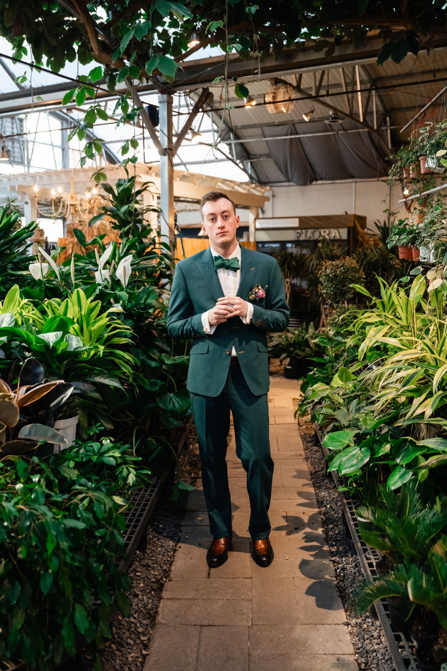 A person in a green suit stands elegantly in a lush greenhouse walkway surrounded by tropical plants and foliage.