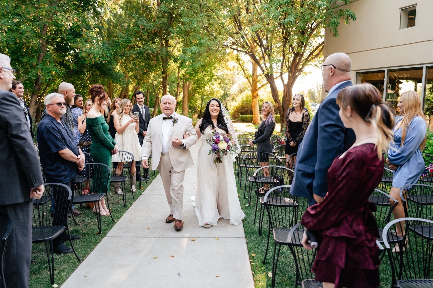 An outdoor wedding ceremony on a garden path with guests seated on metal chairs as golden sunlight filters through trees.