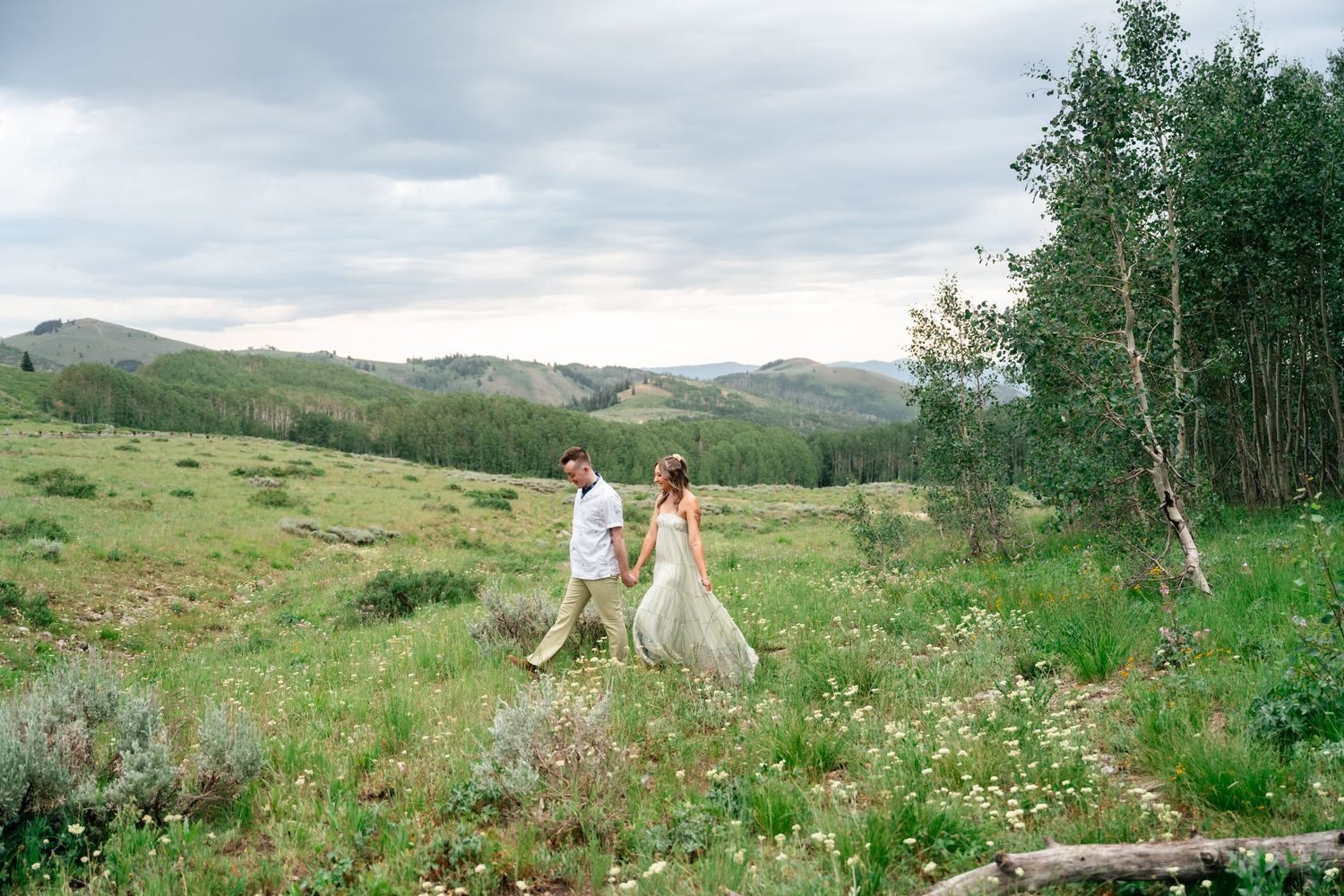Engaged couple walk hand in hand across a green mountain meadow with wildflowers and aspen trees in the background. Park City engagement