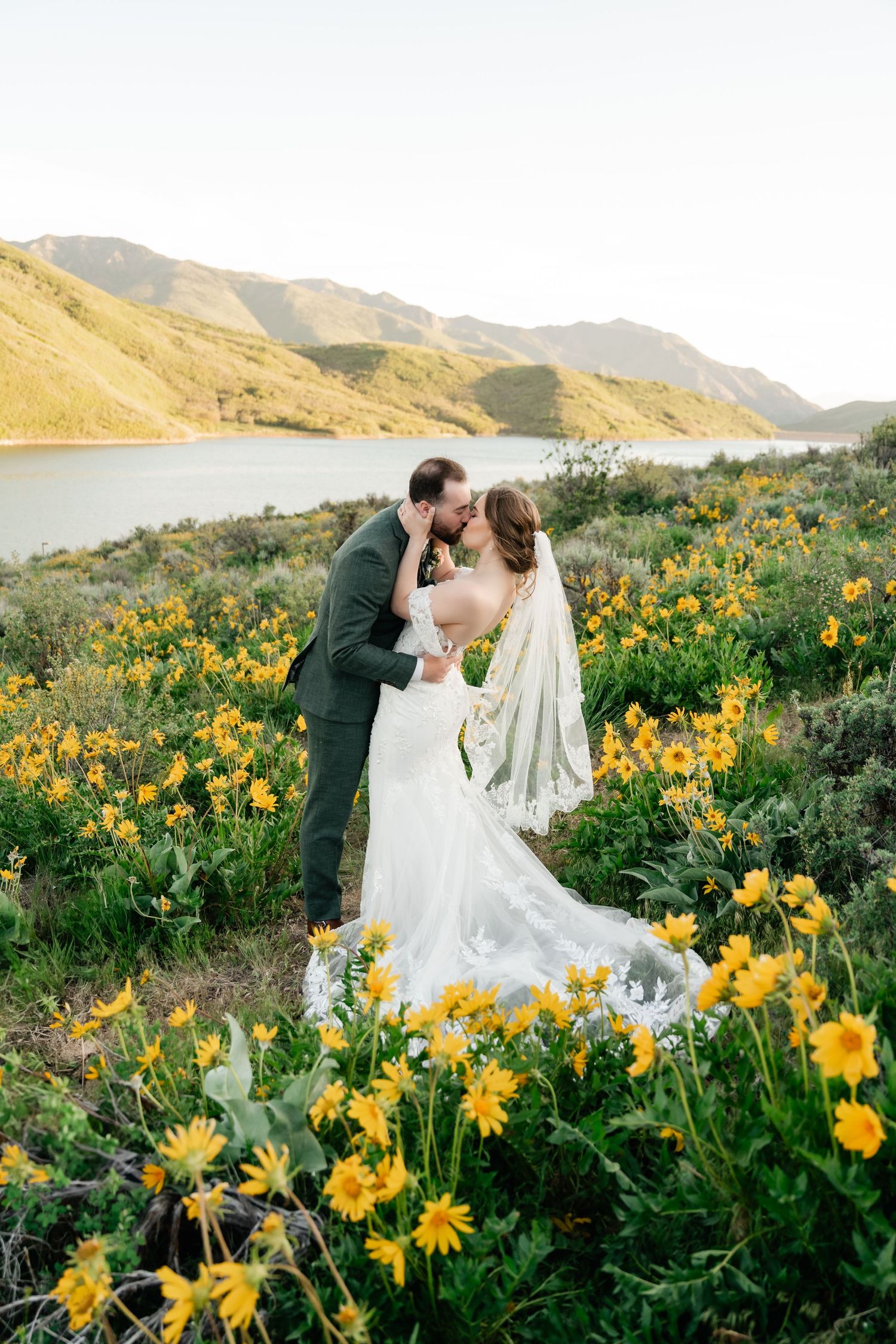 Couple shares romantic moment among yellow wildflowers with mountain lake in background.