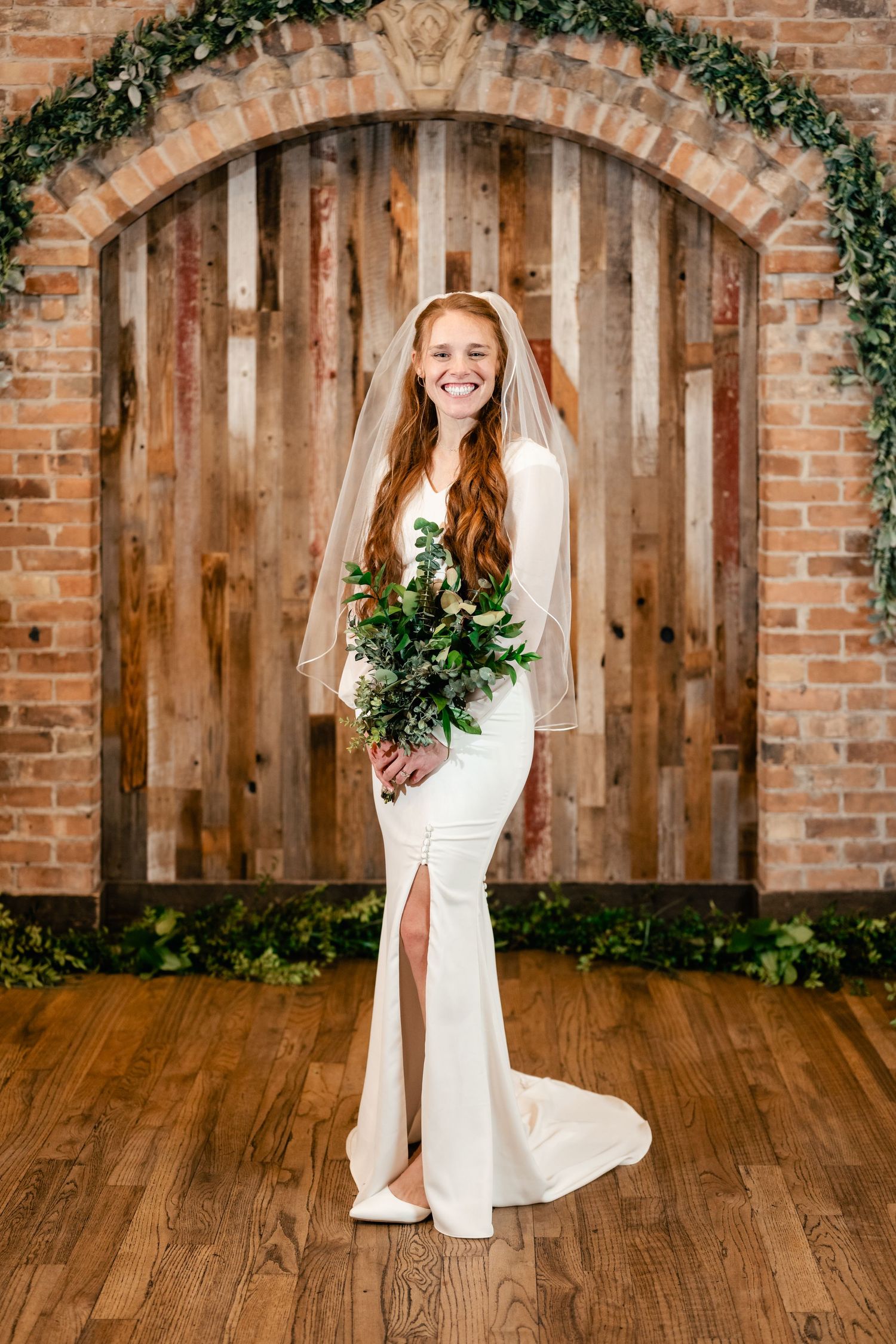 A bride in a fitted white wedding dress with slit holds greenery bouquet against rustic wood and brick archway.
