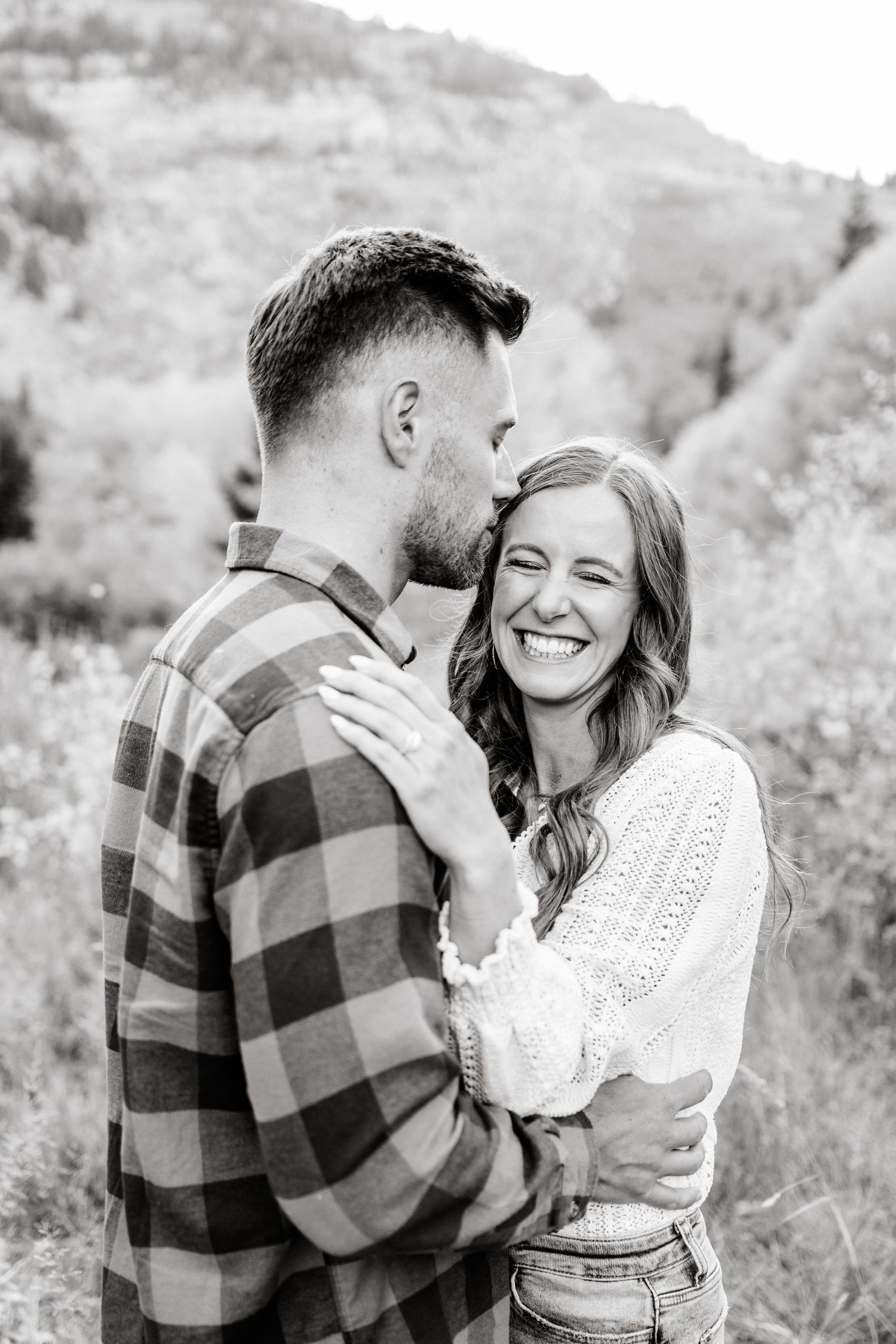A romantic couple shares a tender moment outdoors in a black and white photo against a mountainous backdrop.