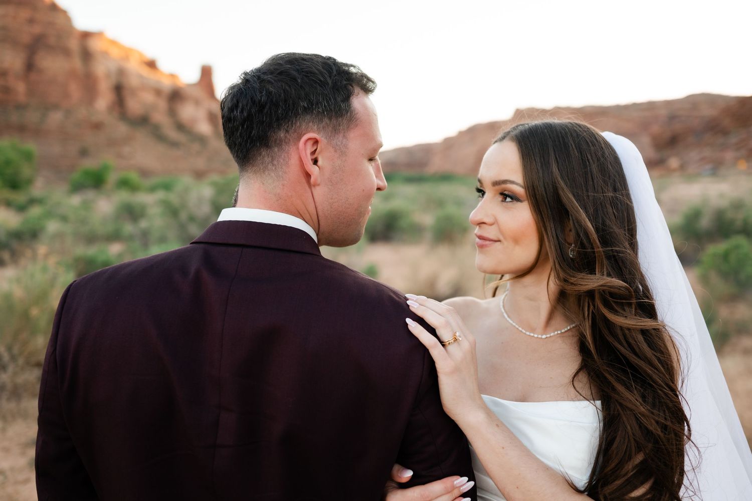 Bride in white gown and veil gazes at groom in burgundy suit against red rock desert backdrop.
