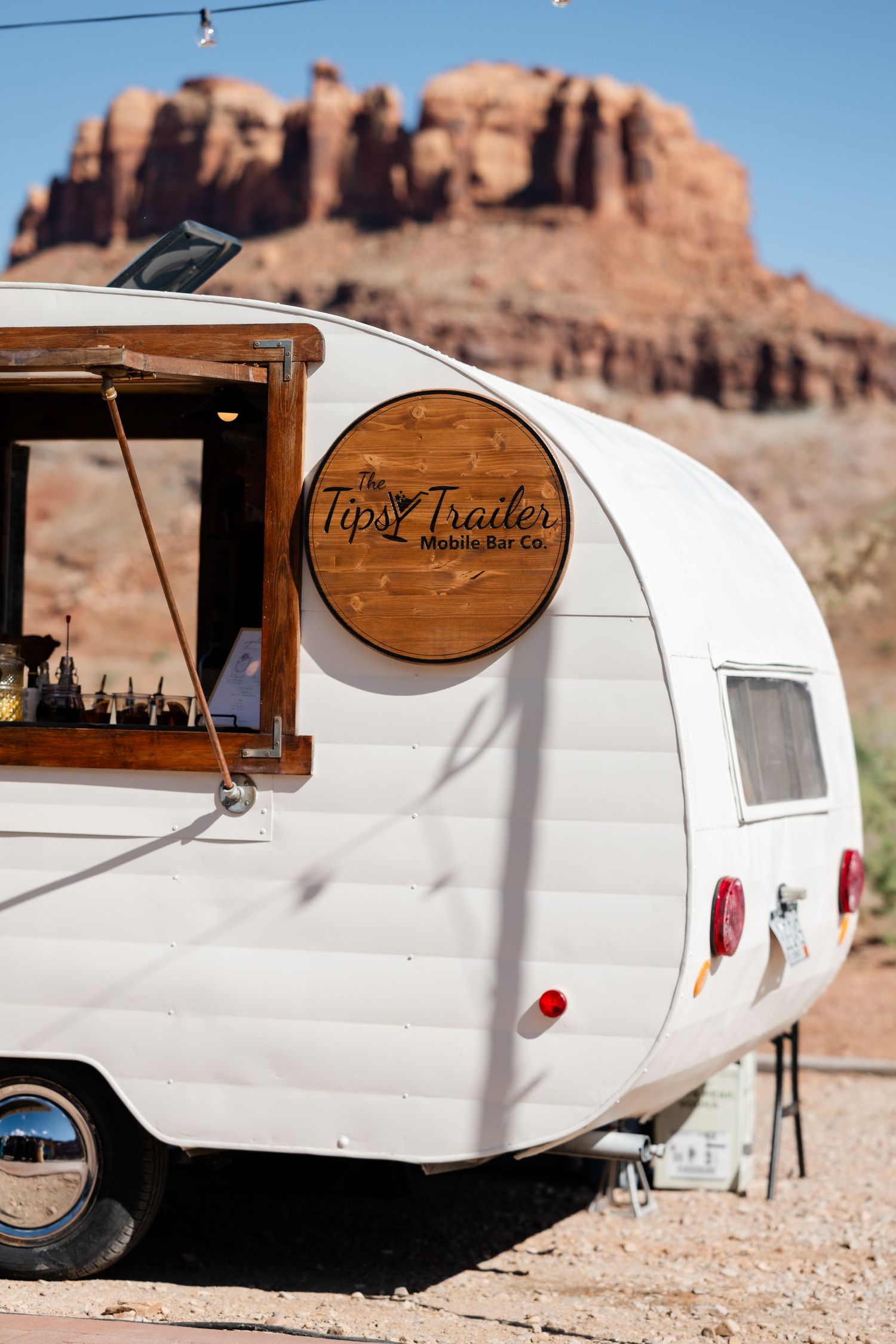 White vintage mobile bar trailer with The Tipsy Trailer sign parked against red rock desert landscape.