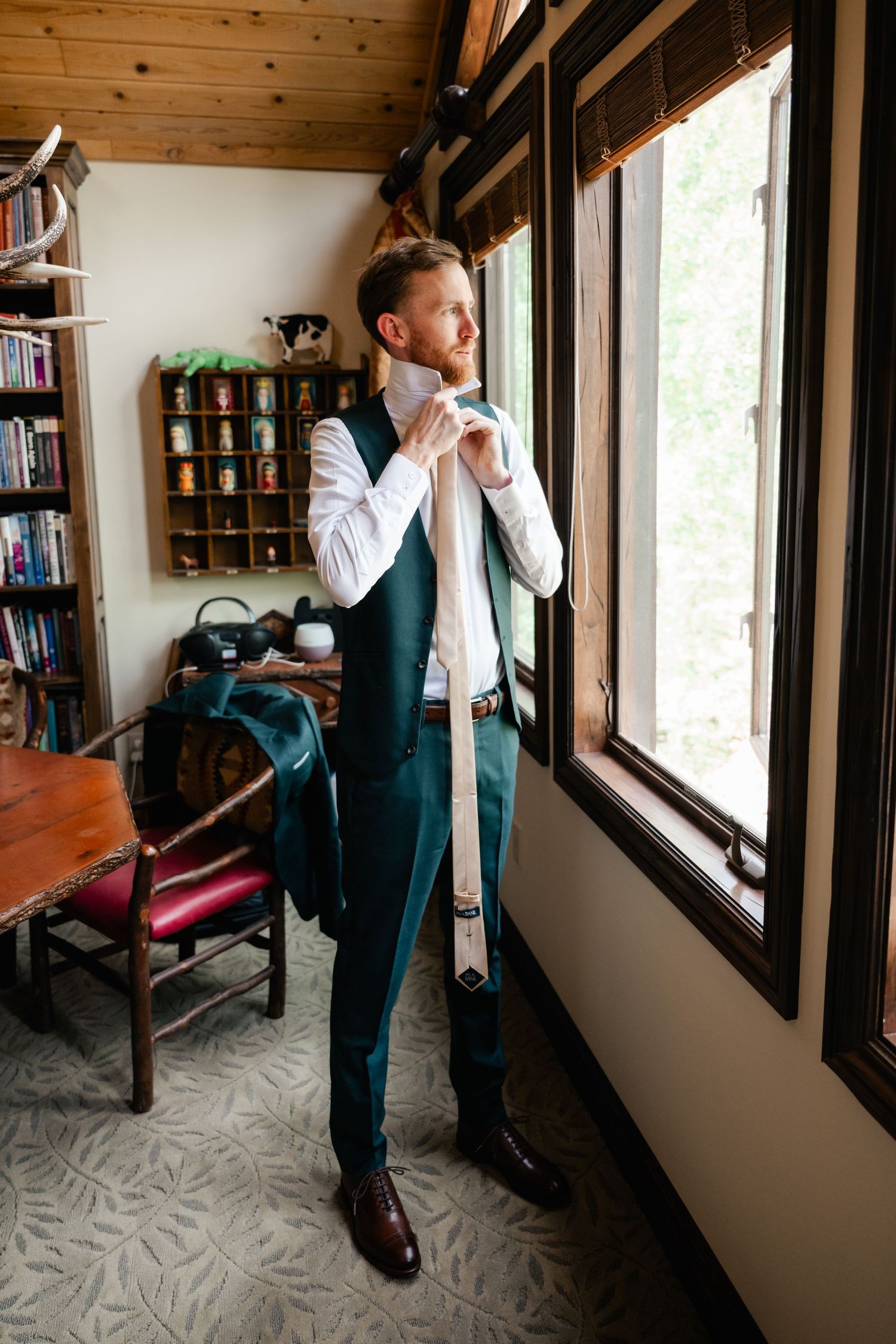 Person adjusting tie while standing by window in rustic room with wooden ceiling and bookshelves.