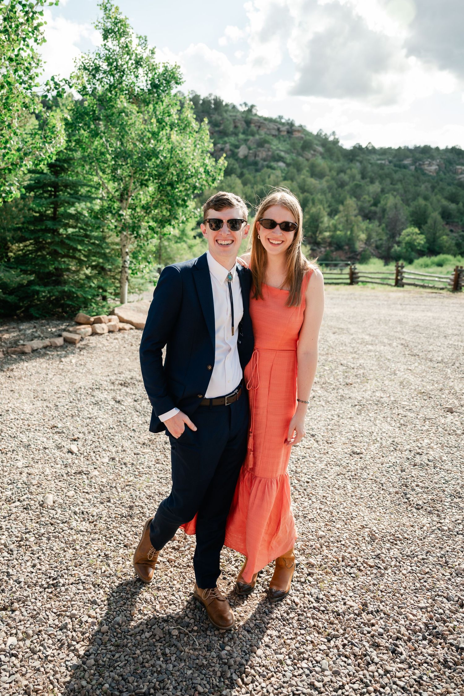 A couple in formal attire poses on a gravel path with mountain scenery in the background.