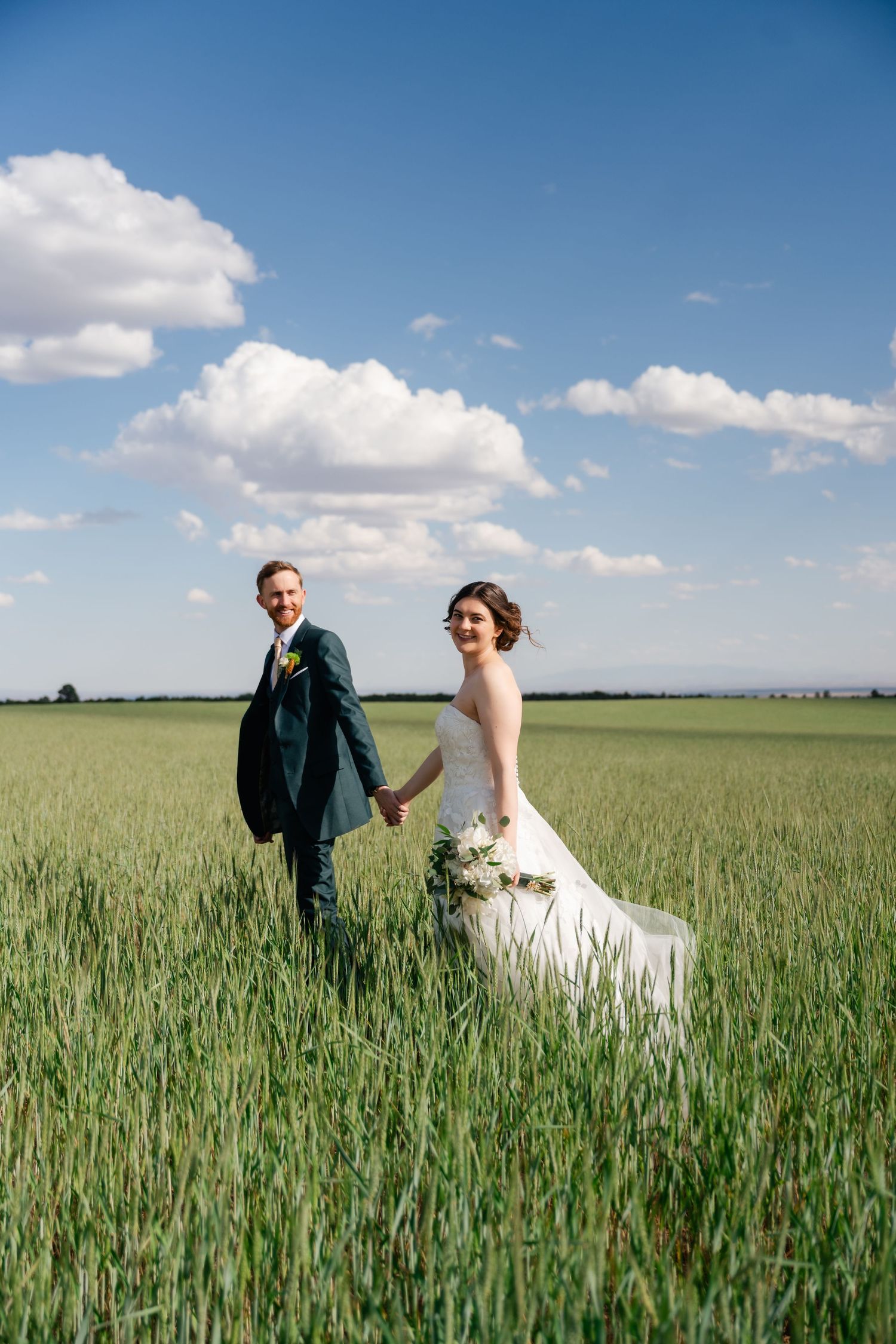 A couple in wedding attire walks through a lush green wheat field under a bright blue sky with white clouds.