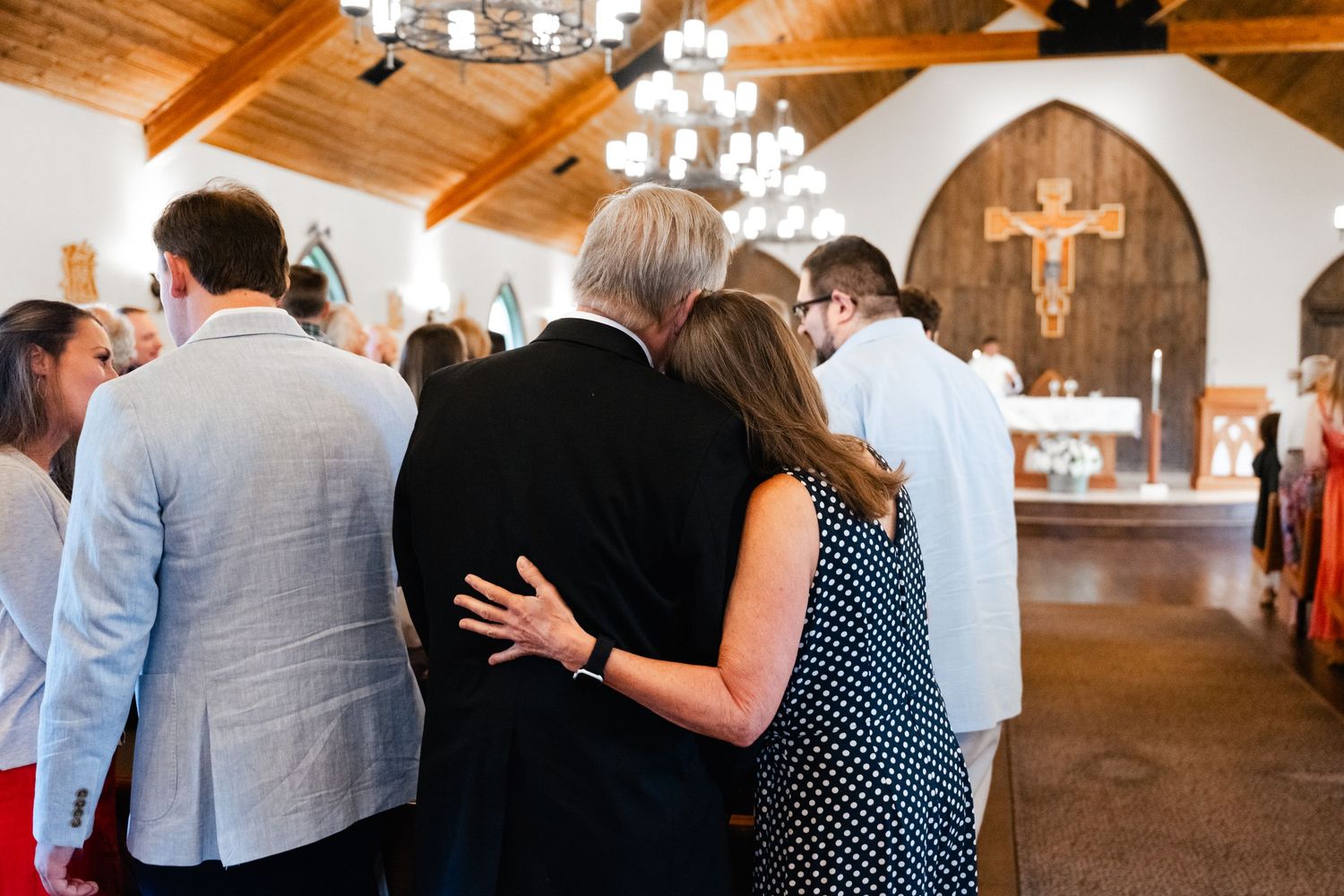 Wedding guests gather in a rustic church venue with wooden ceiling beams and chandeliers.