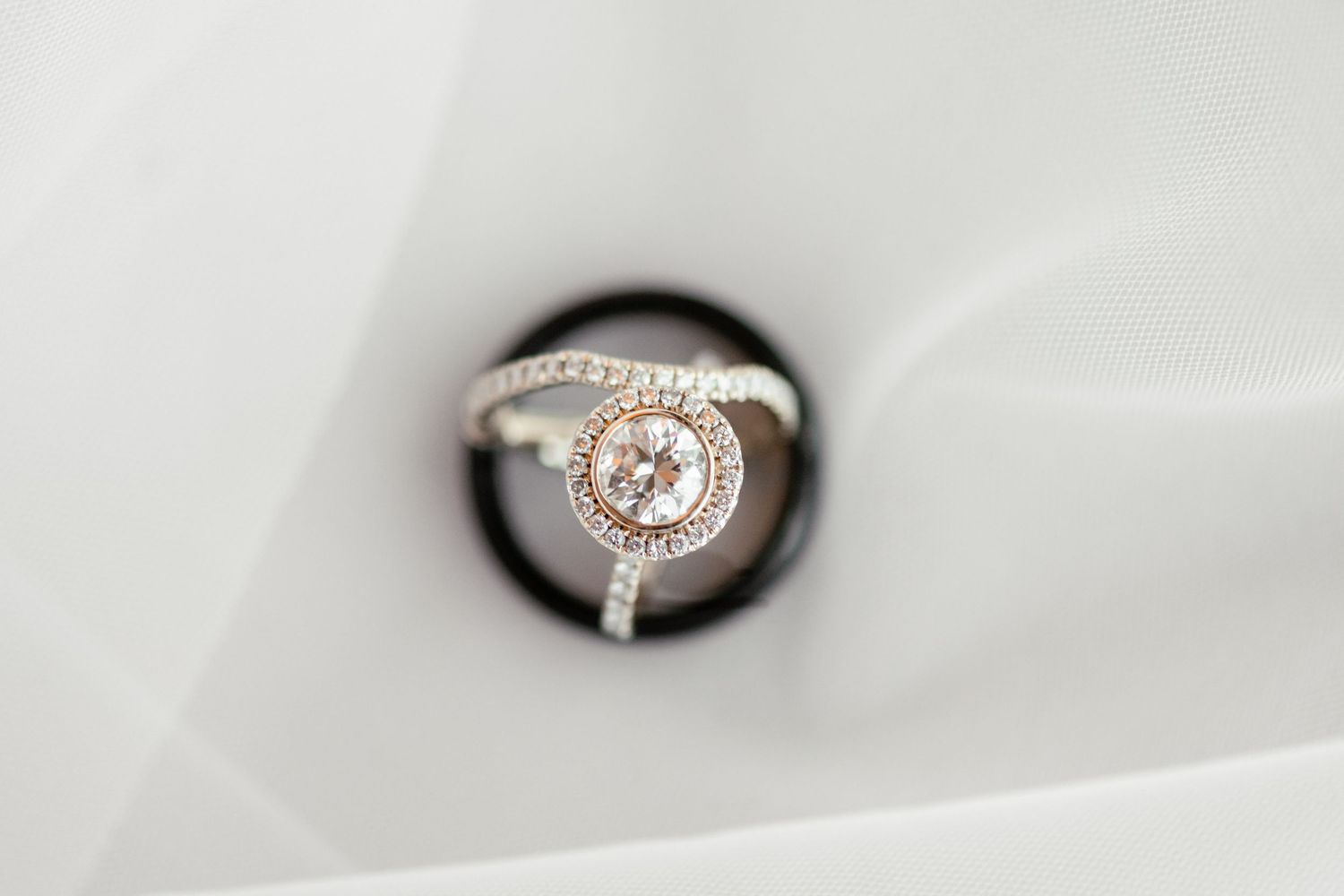 A sparkling diamond engagement ring with halo design photographed through circular opening on white fabric background.