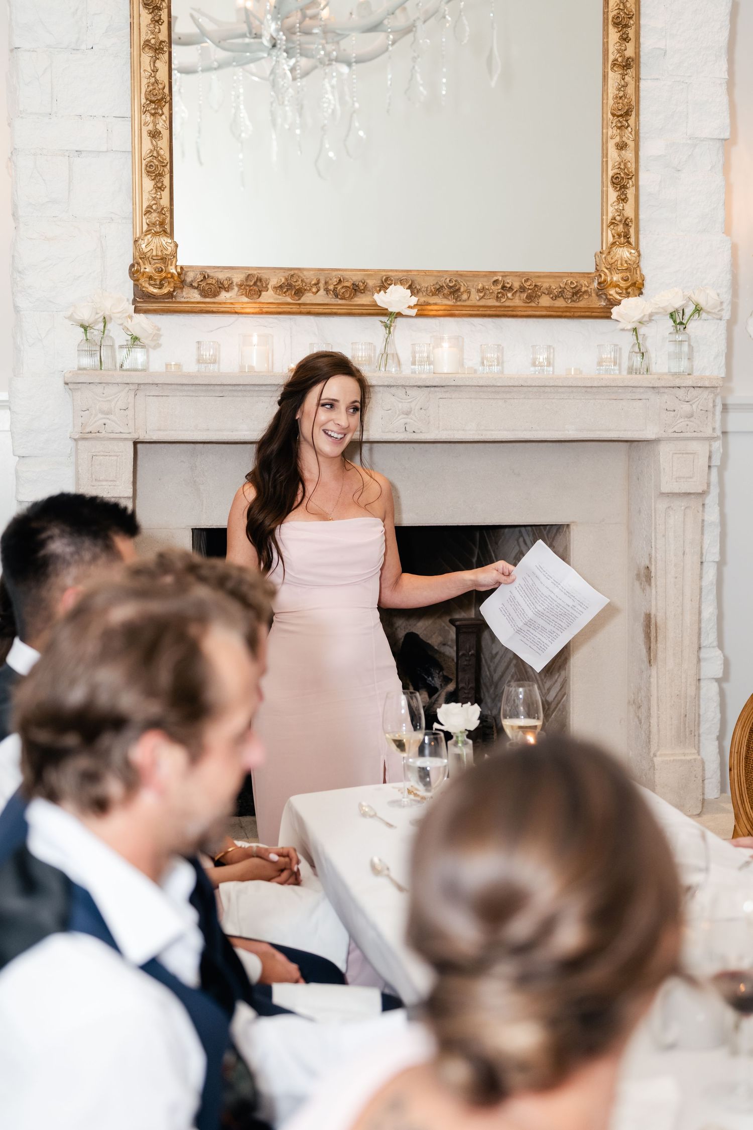 Wedding reception speech being given in an elegant white room with ornate gold mirror and fireplace.