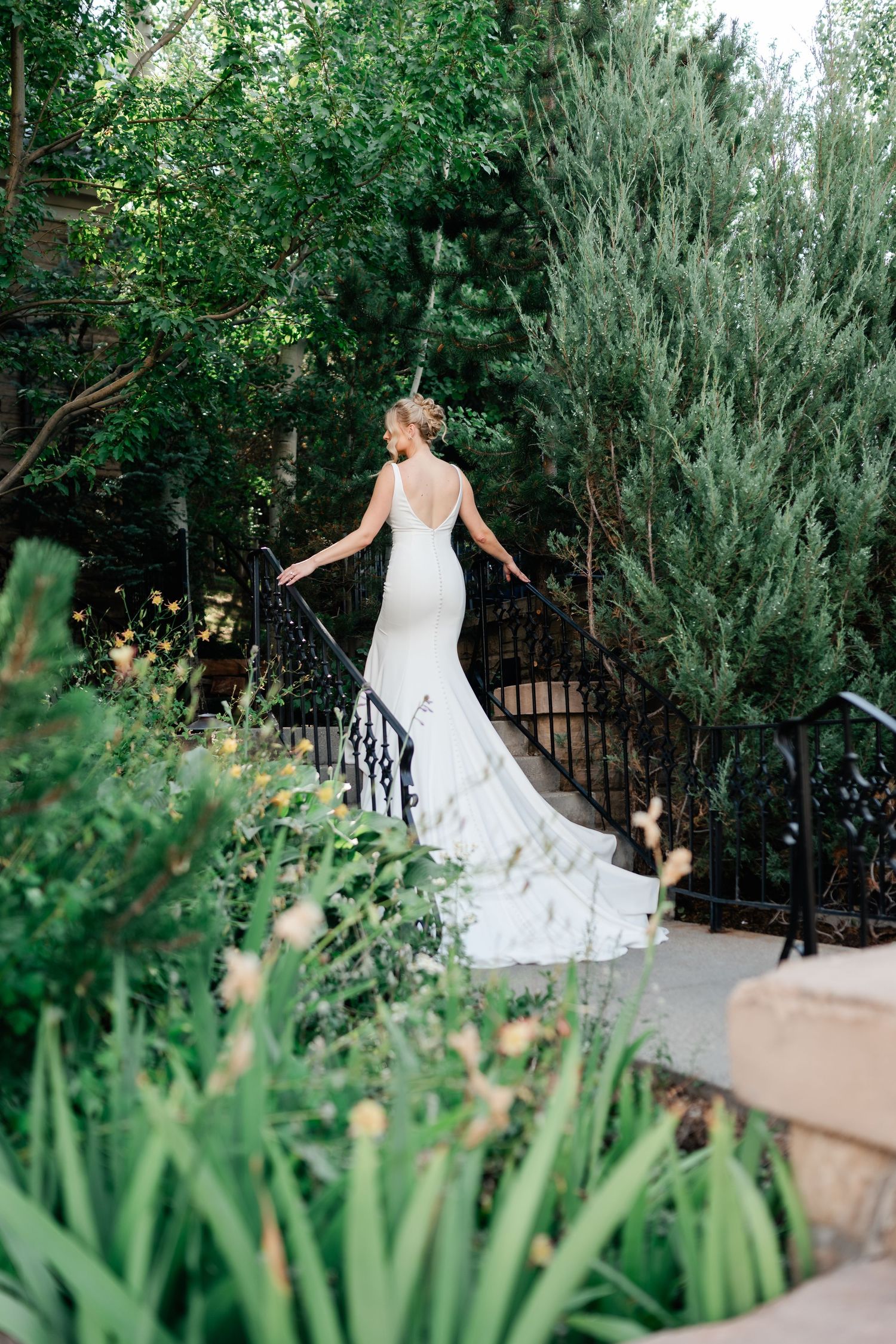 Elegant bride in fitted wedding dress poses on garden path with ornate iron railing.