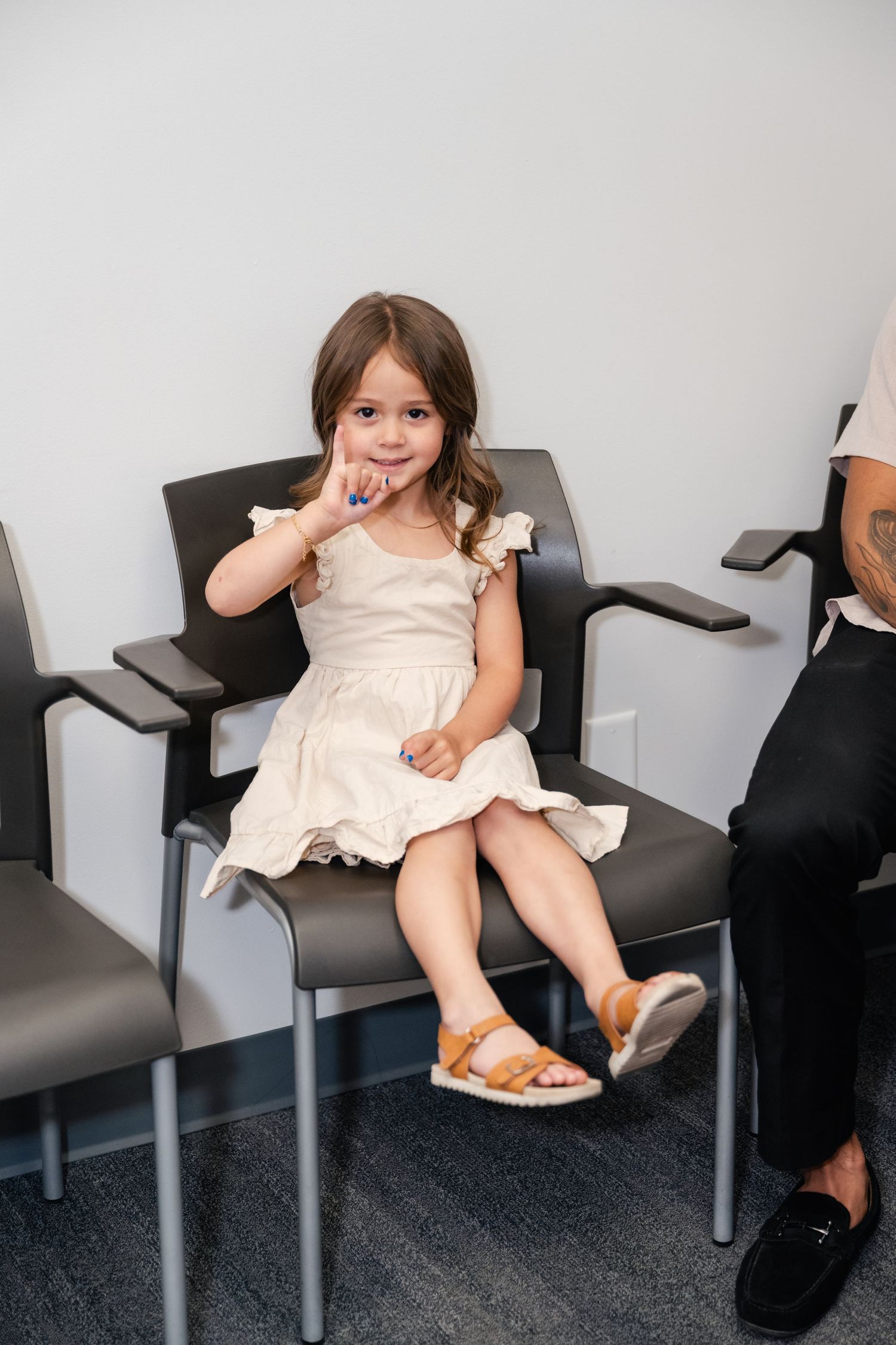 A young child in a cream-colored dress sits cheerfully in a waiting room chair.