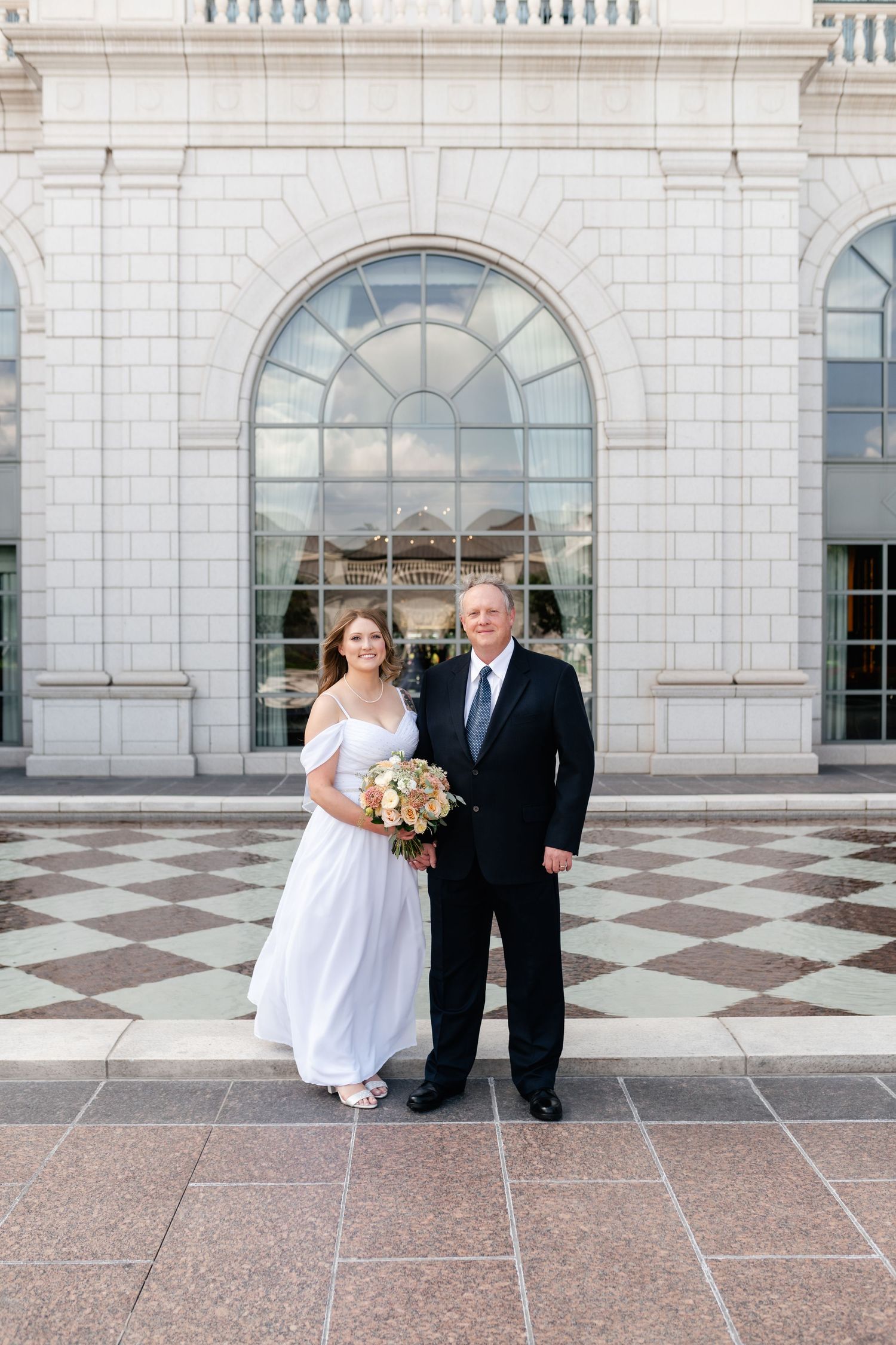 A wedding couple stands on a checkered tile floor in front of an elegant arched window at a grand building entrance.