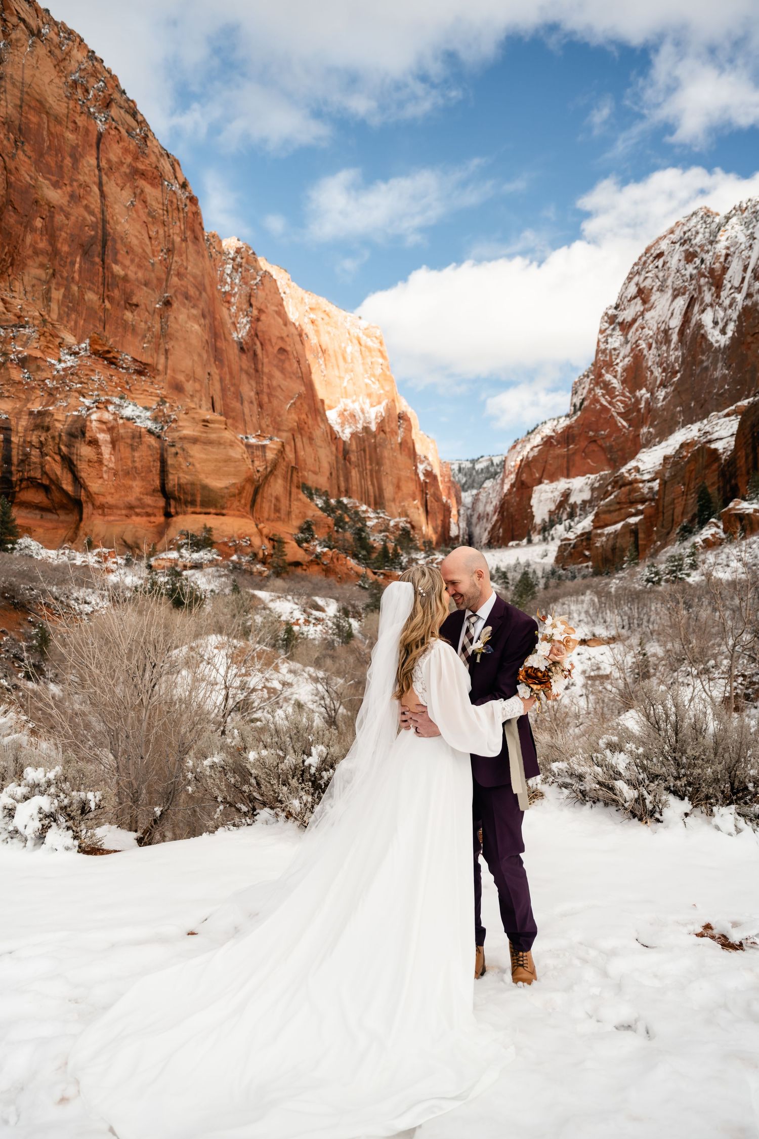 A winter wedding couple embraces against stunning red rock cliffs and snowy landscape in a national park.