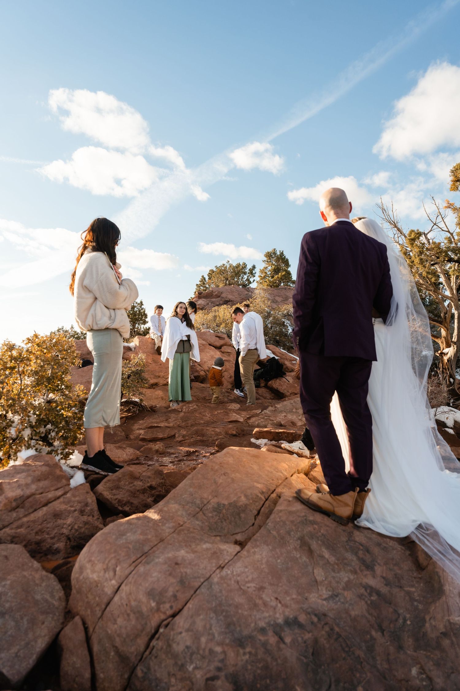Wedding party stands on red rock formations during an outdoor ceremony against a bright blue desert sky.
