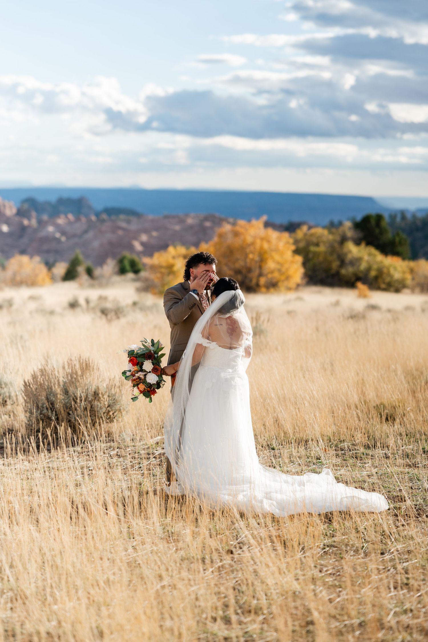 A romantic wedding couple embraces in a golden field with mountains and autumn trees in the background.